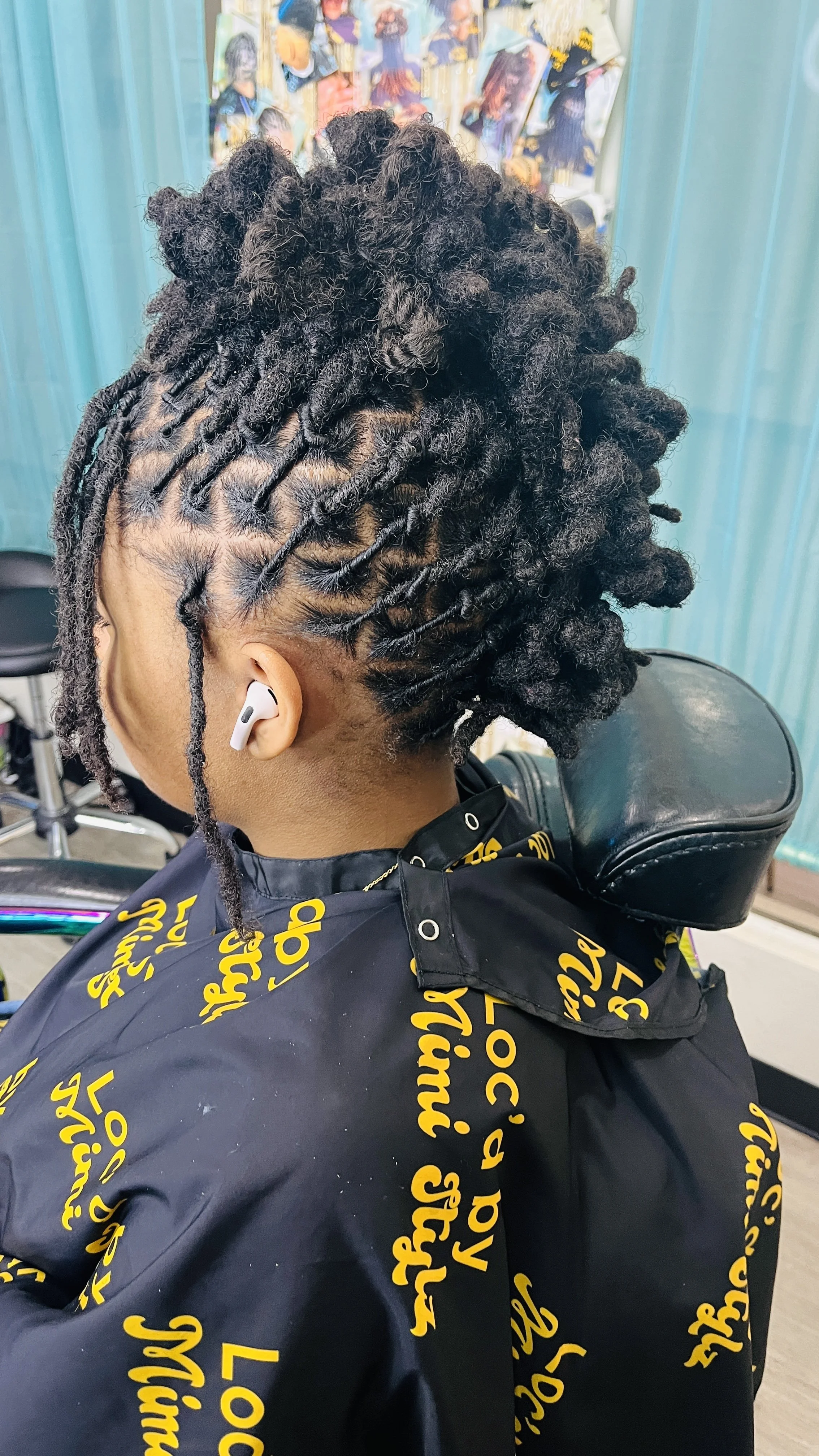 Close-up of a person with short dreadlocks styled into an updo, sitting in a salon chair with a black cape that has yellow script lettering. The background includes turquoise walls and colorful artwork.