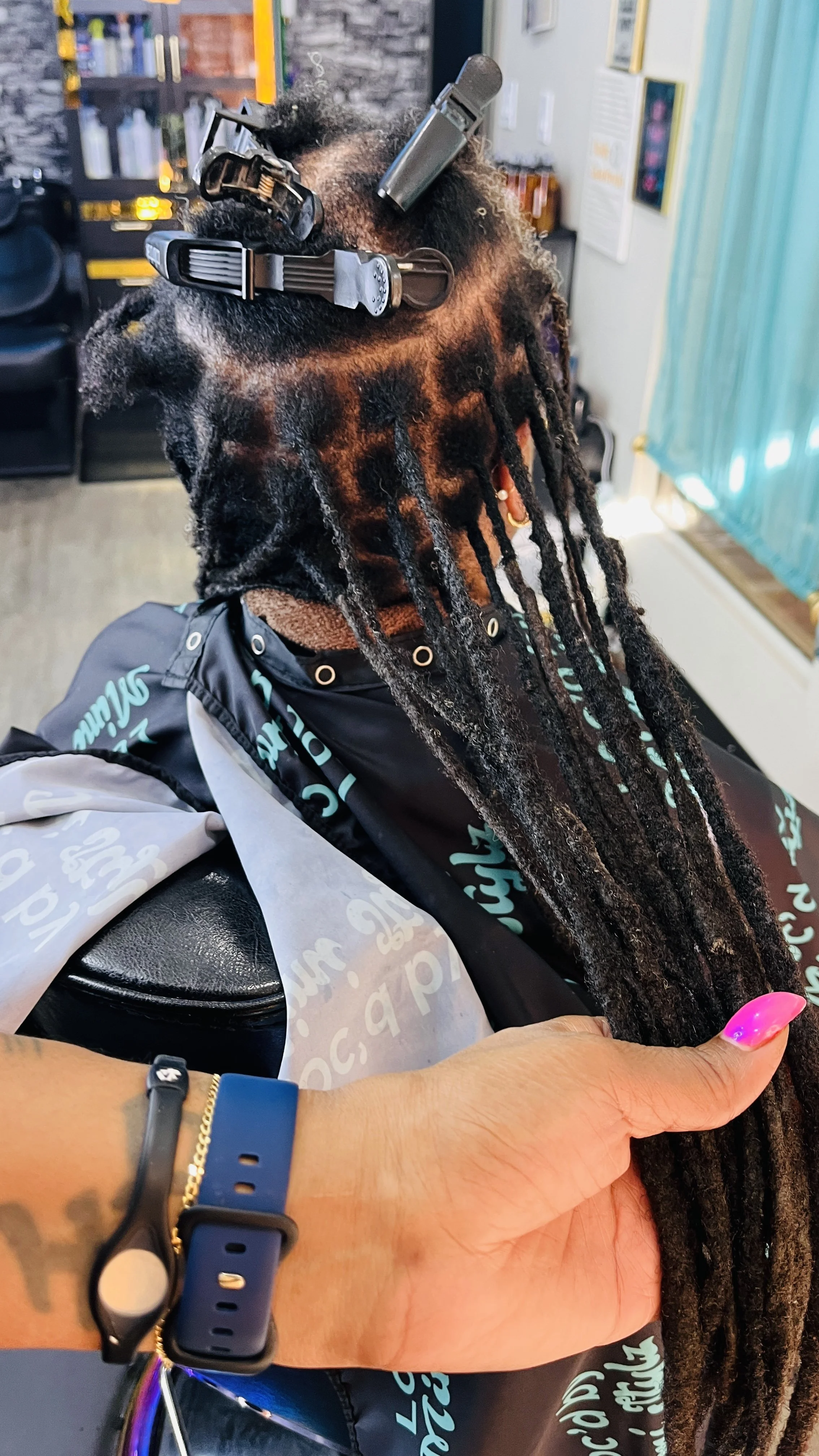 A person with dreadlocks getting a haircut at a salon, with hair clips and a razor on their hair, and a stylist's hand holding their head.