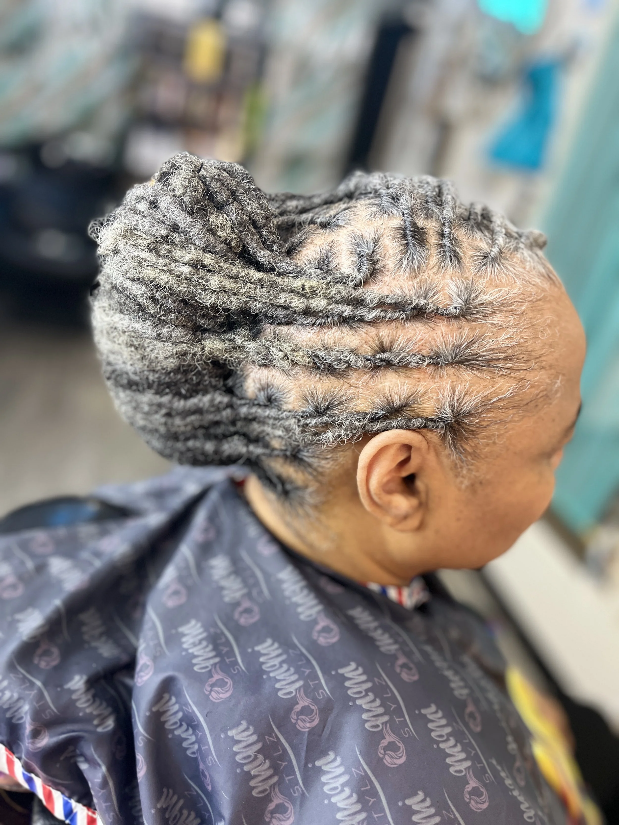 Person with neatly styled gray twists or locks in their hair, wearing a dark patterned cape typical of a salon, getting a hair treatment or style at a salon.