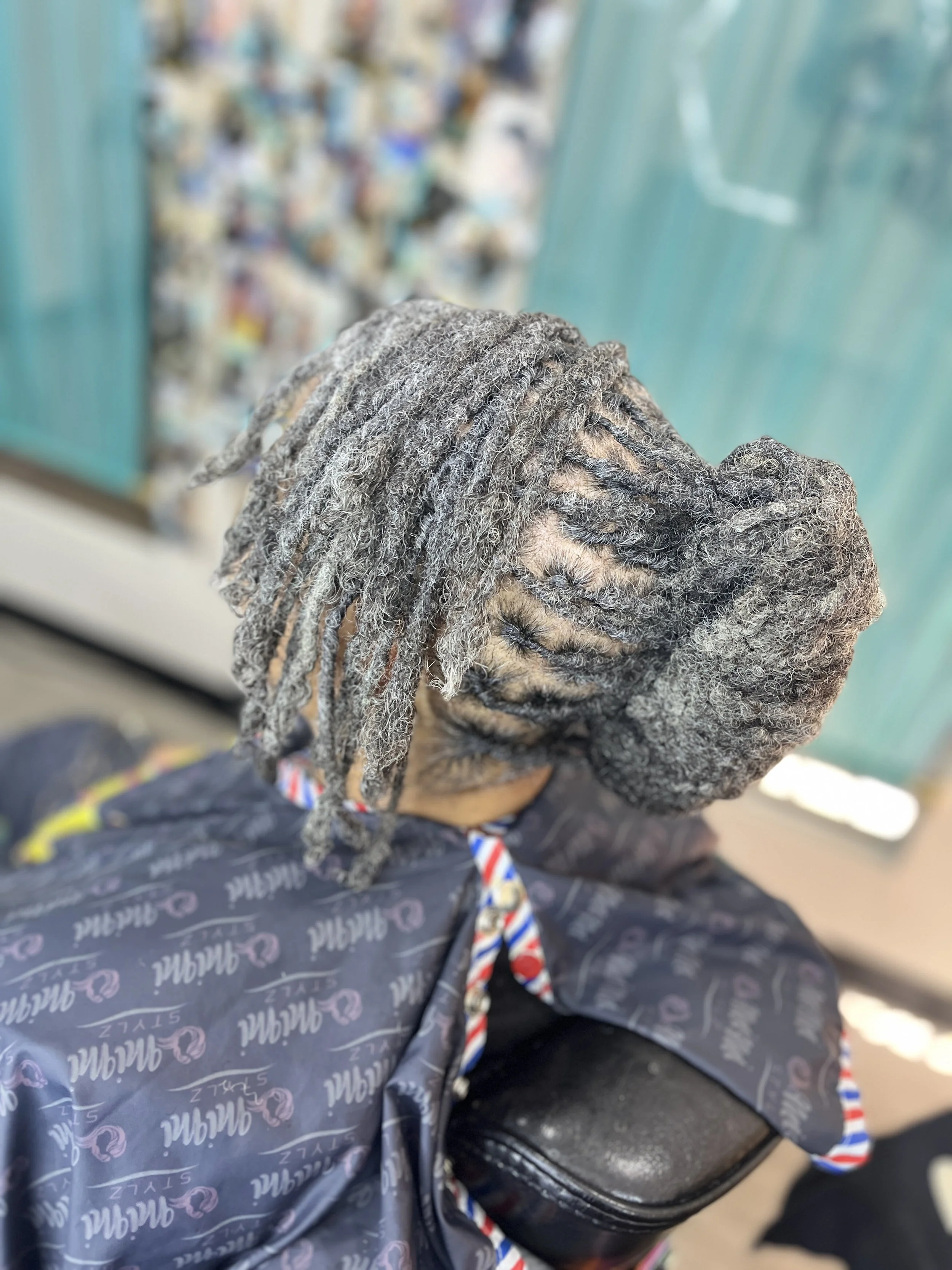 Close-up of person's head with gray dreadlocks, wearing a jacket with a patterned logo, sitting on a black chair, with a blurred background of a vending machine.