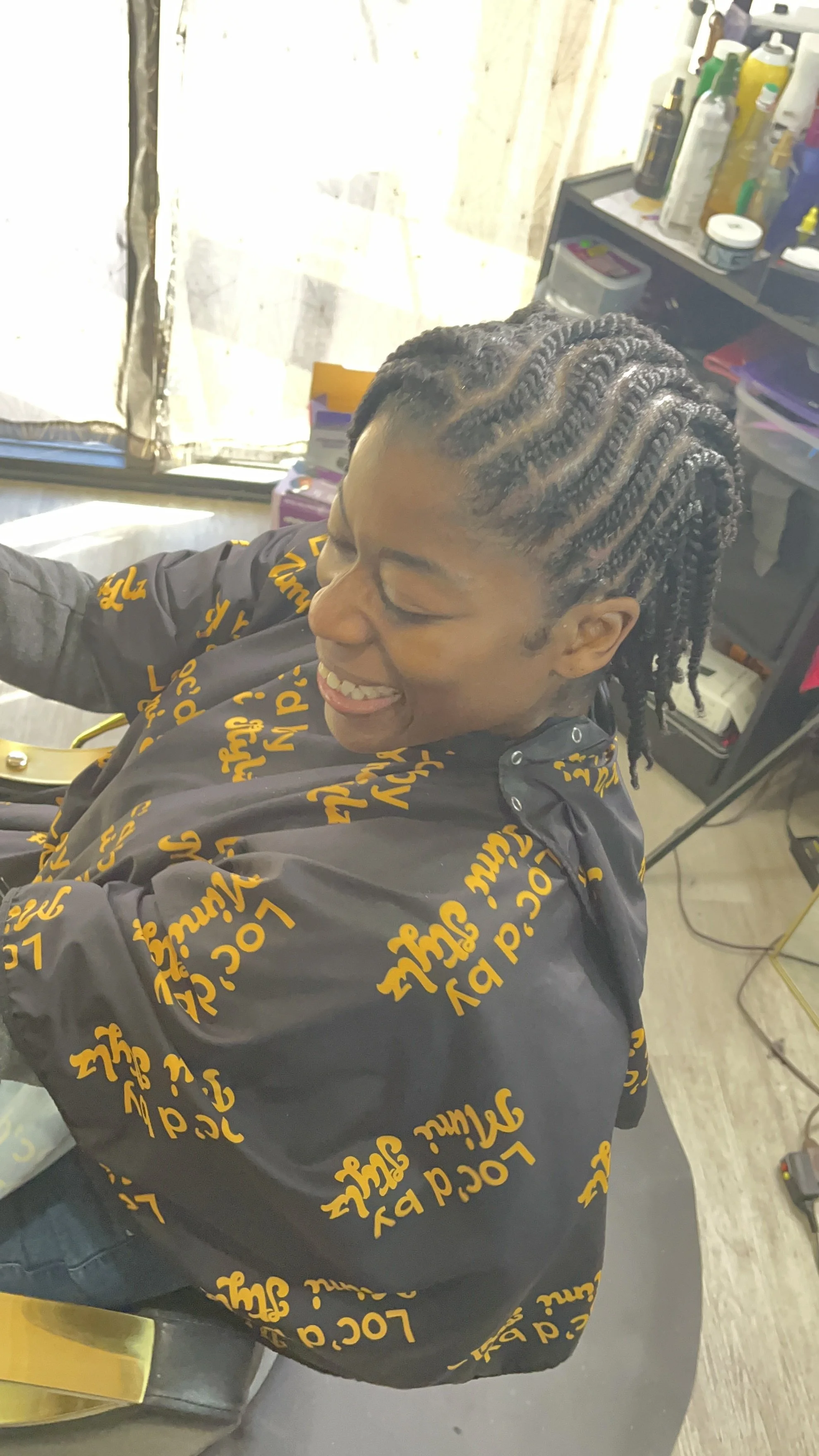 A woman with styled dreadlocks smiling while sitting in a hair salon chair, wearing a black cape with yellow writing, surrounded by hair products and salon equipment.