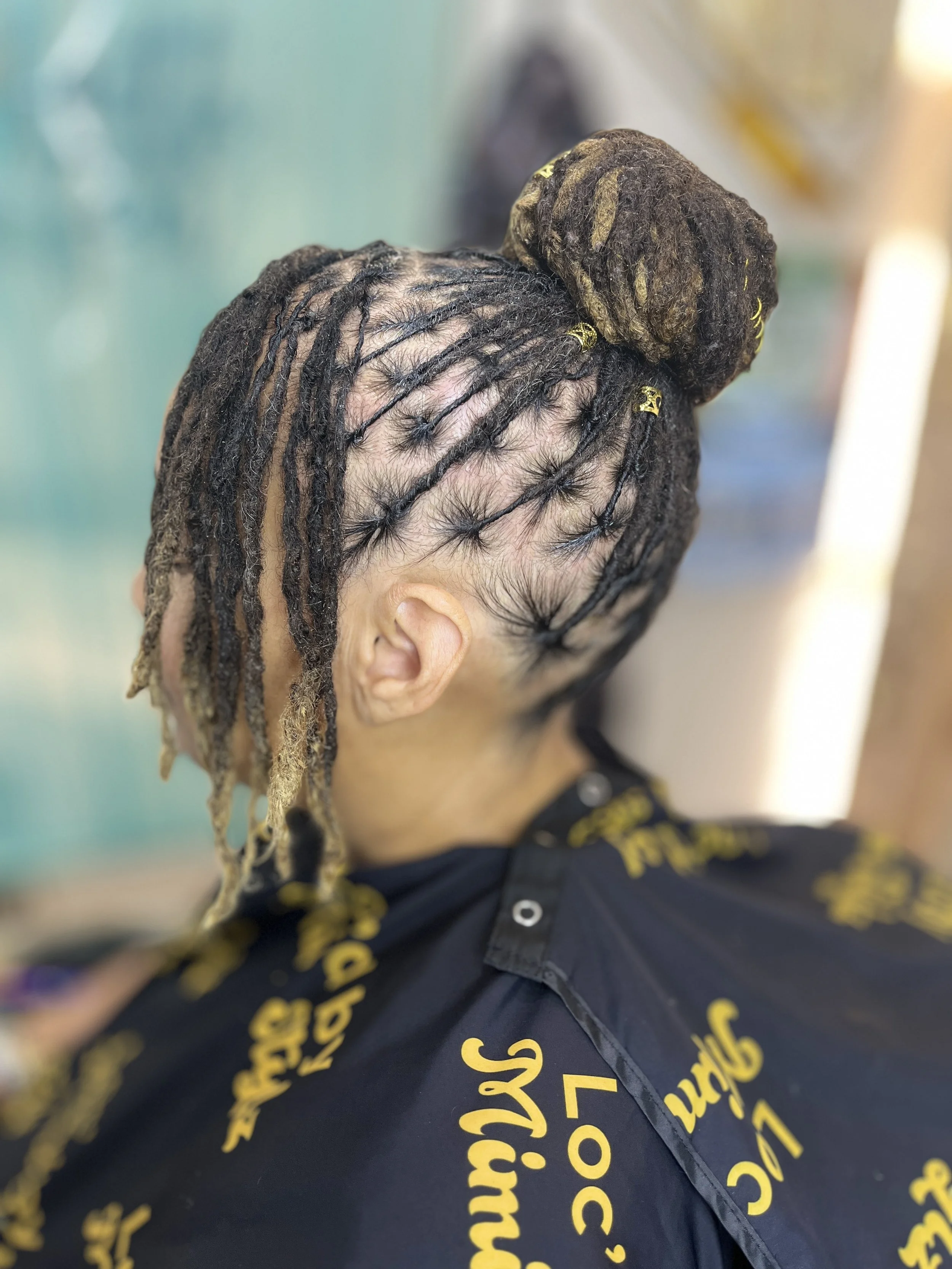 Close-up of a person's head with dreadlocks styled in a bun, wearing a black shirt with yellow text, in an indoor setting.