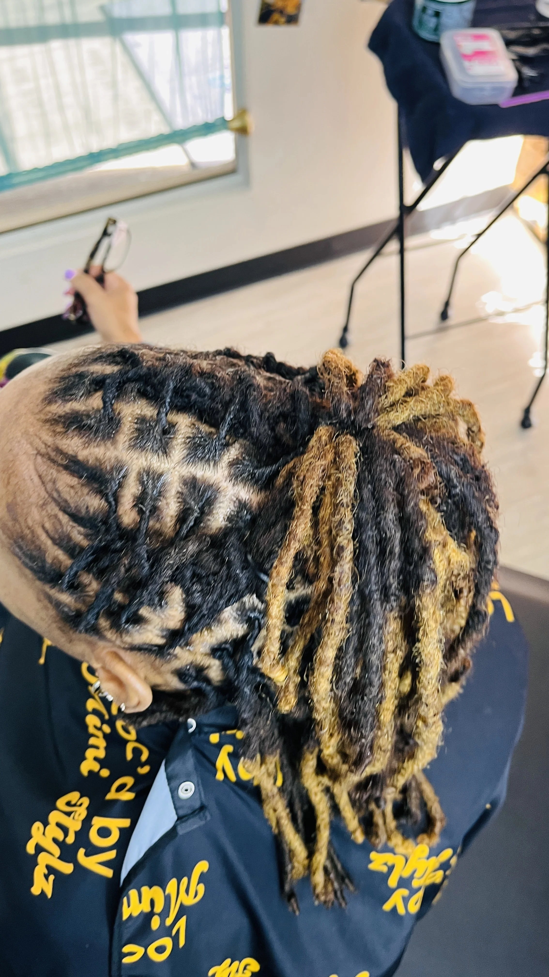 Person with styled dreadlocks sitting in a room near a window, wearing a dark jacket with yellow text, holding a phone, with a table and some items on it in the background.