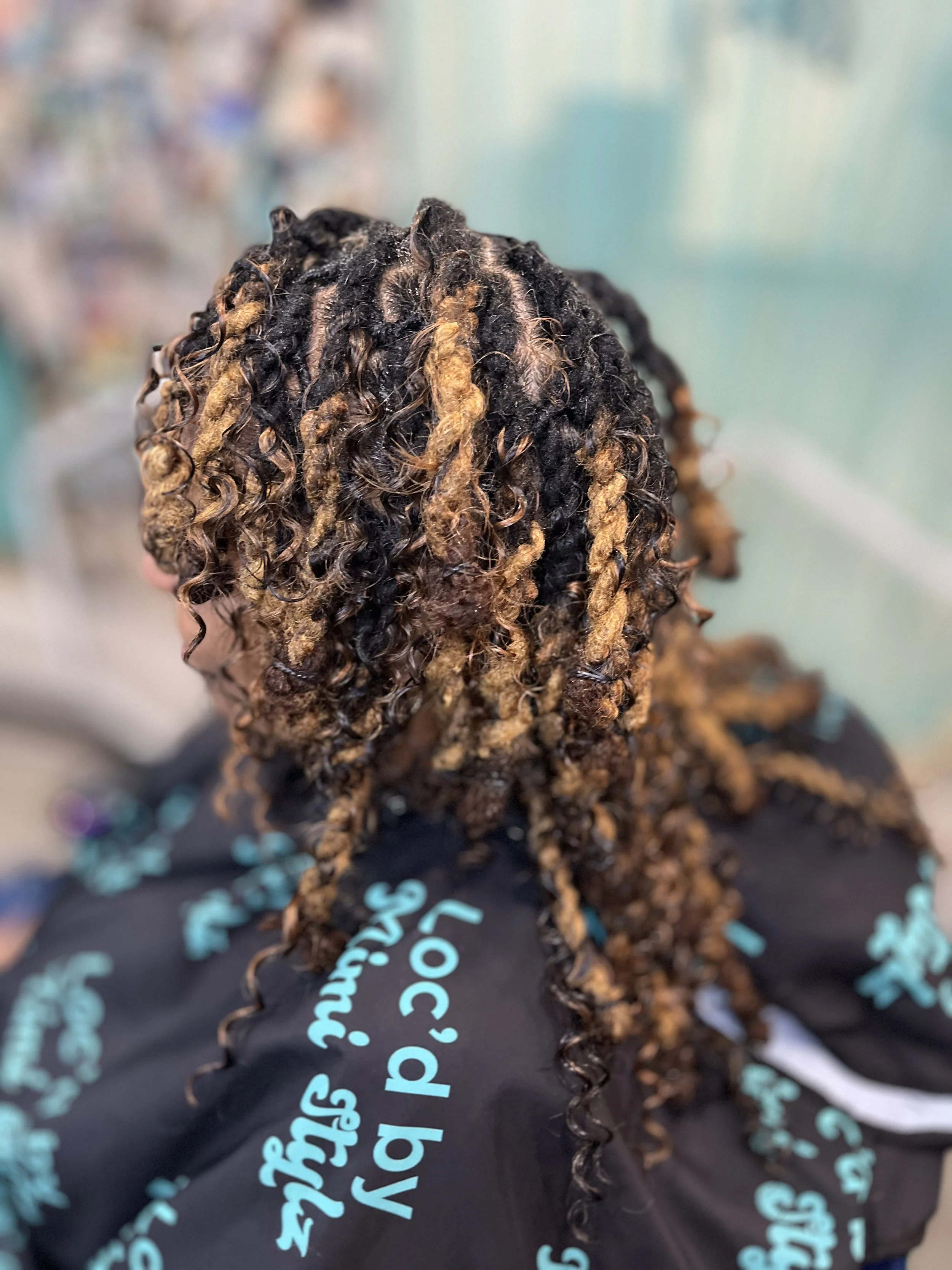 Close-up view of a person's head with styled dreadlocks, with some locks dyed a light brown color, and the person is wearing a black shirt with blue text.
