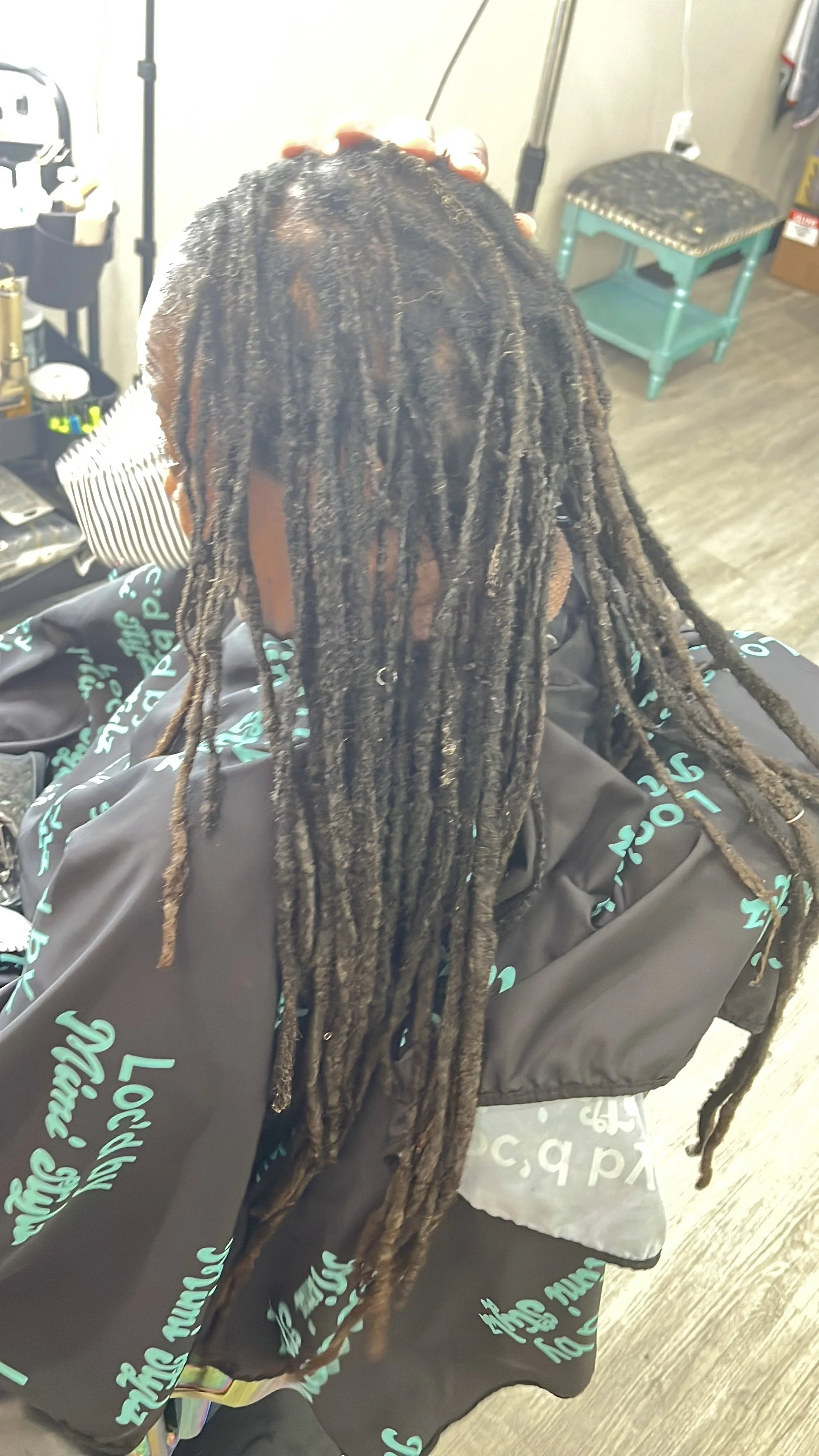 Person with long dreadlocks sitting in a salon chair, facing away from the camera, with a hand resting on their head.