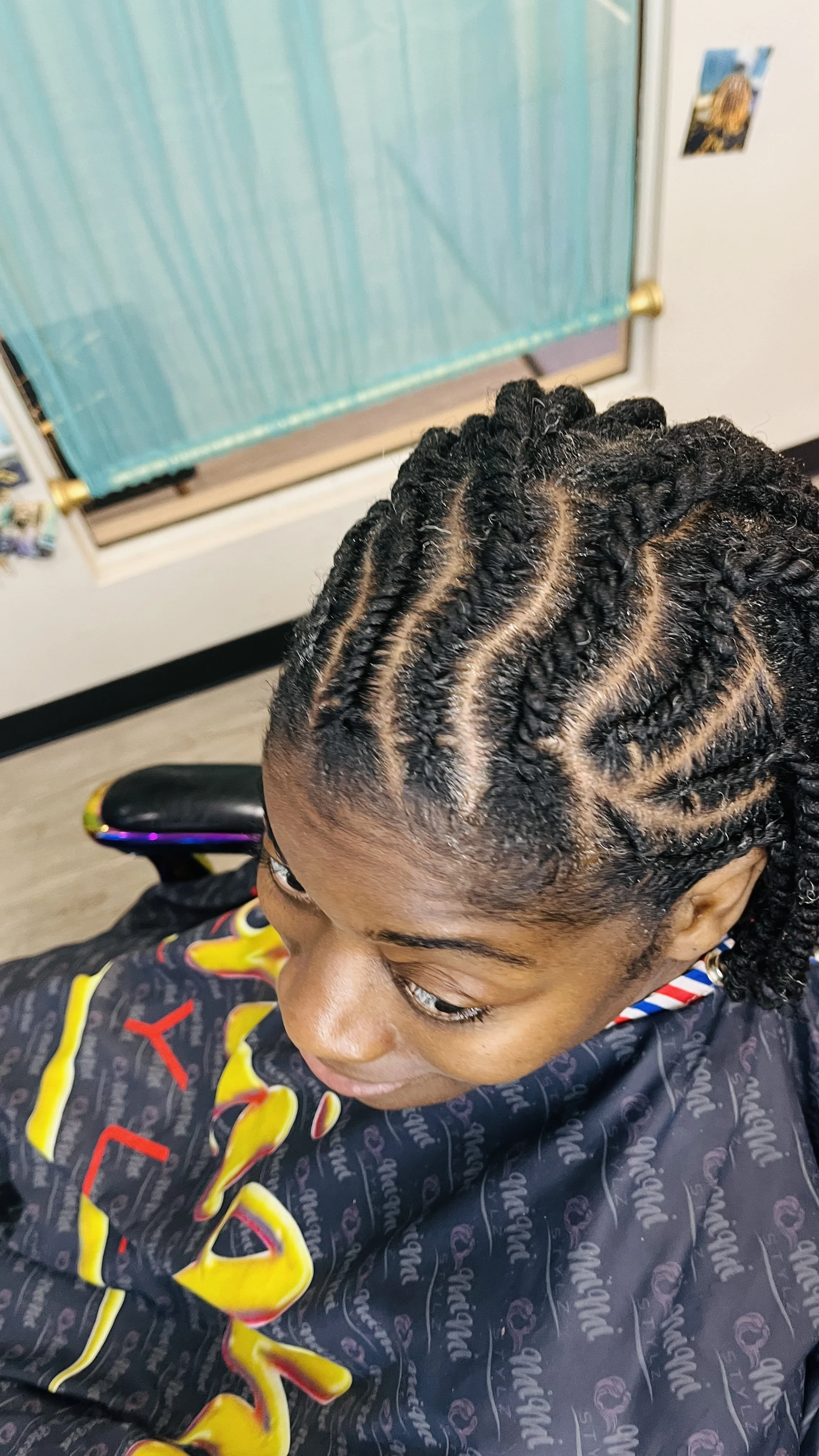Close-up of a person with neatly styled black braided hair with a patterned cape, sitting in a chair in a salon, with a spray bottle and a table with hairdressing tools in the background.