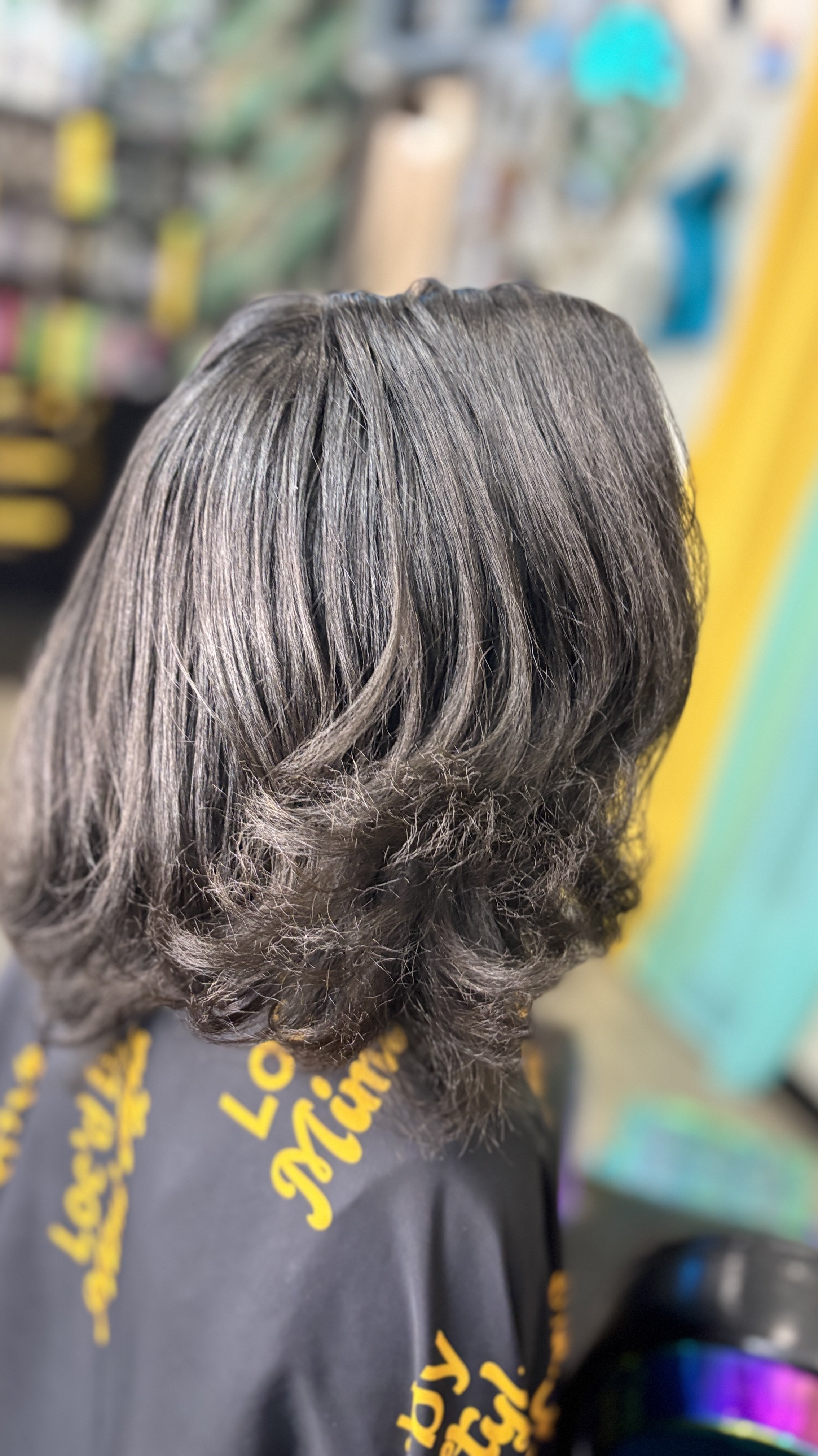 Back of a person's head with black hair, silk press with light waves, in The Thairapy Suite with colorful blurred background.