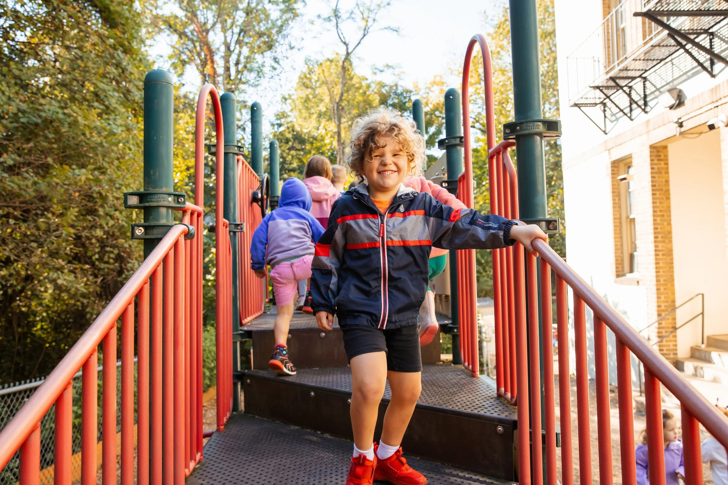 Kids playing on a playground structure, with a smiling boy in the front, and other students climbing the play structure in the background, during a sunny day.