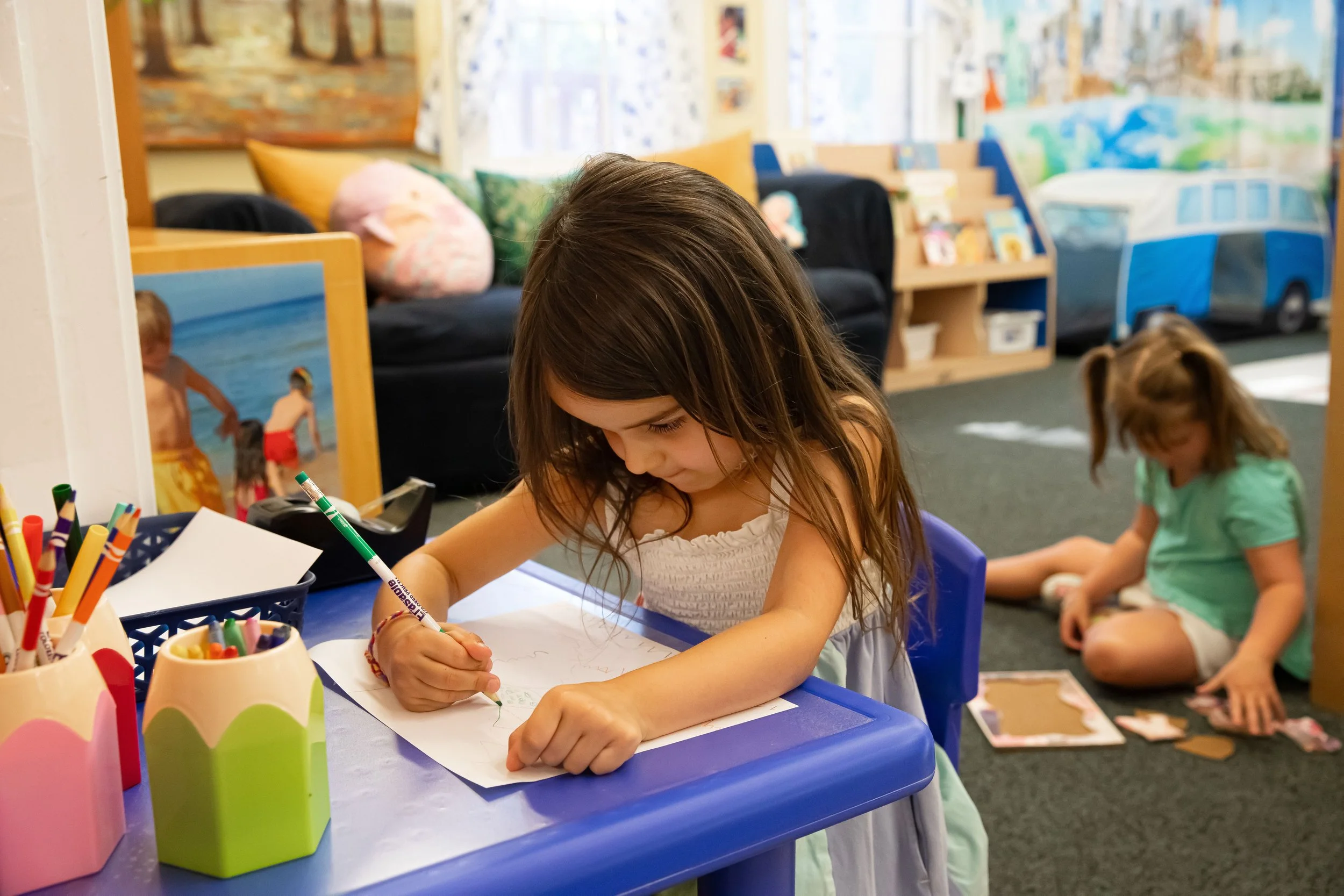One student sits at a blue table, drawing on paper with a green marker, surrounded by colored pencils. In the background, another student is sitting on the carpet playing with toys.