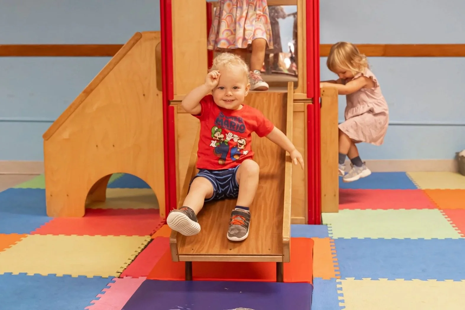 Two young children playing on an indoor wooden play structure with a slide. The boy in the foreground is smiling while going down the slide. A girl in the background is climbing up the play structure.