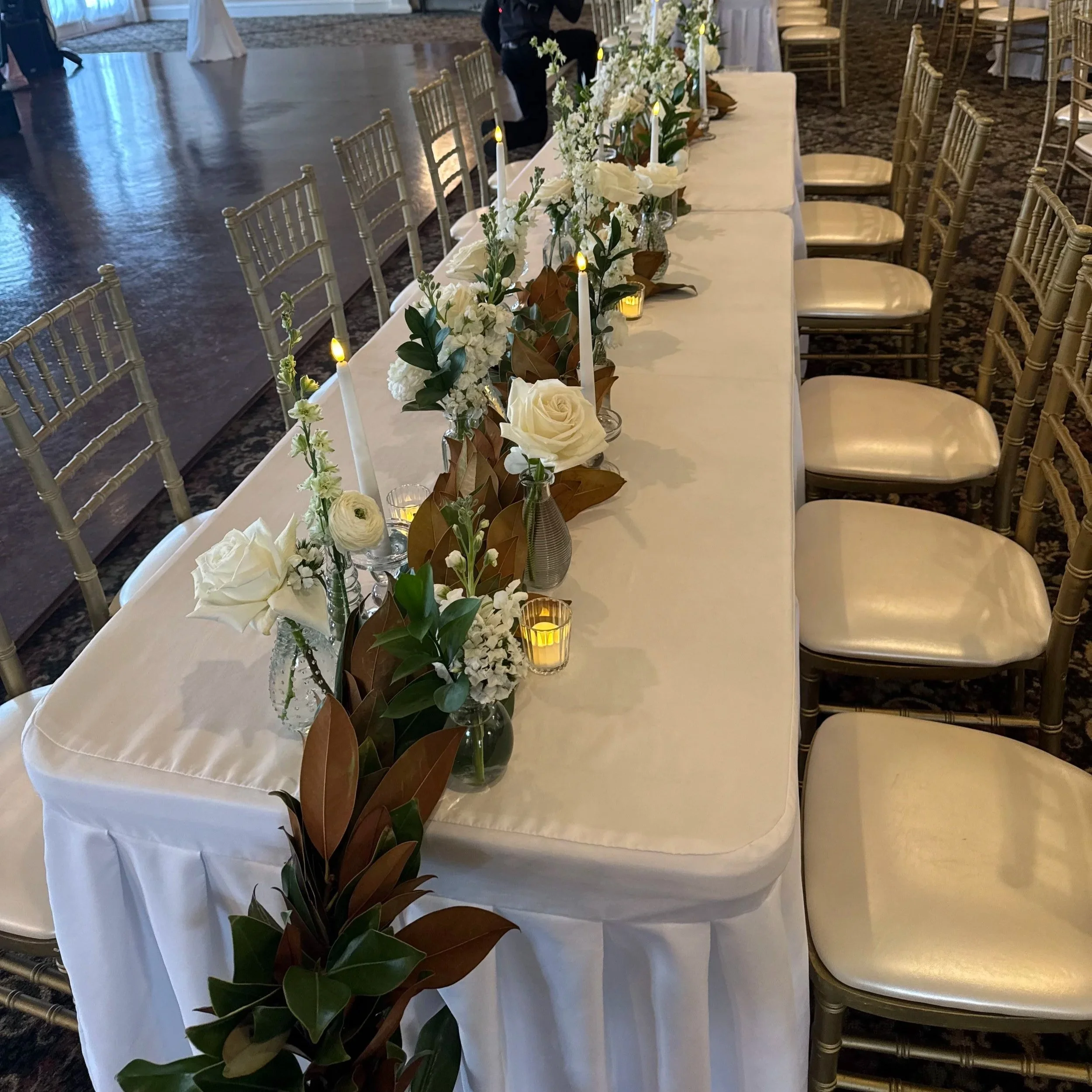 A lavishly decorated room with warm lighting, floral arrangements, candles, draped curtains, vintage furniture, and numerous candles and rose petals on the floor, creating an elegant and romantic ambiance.