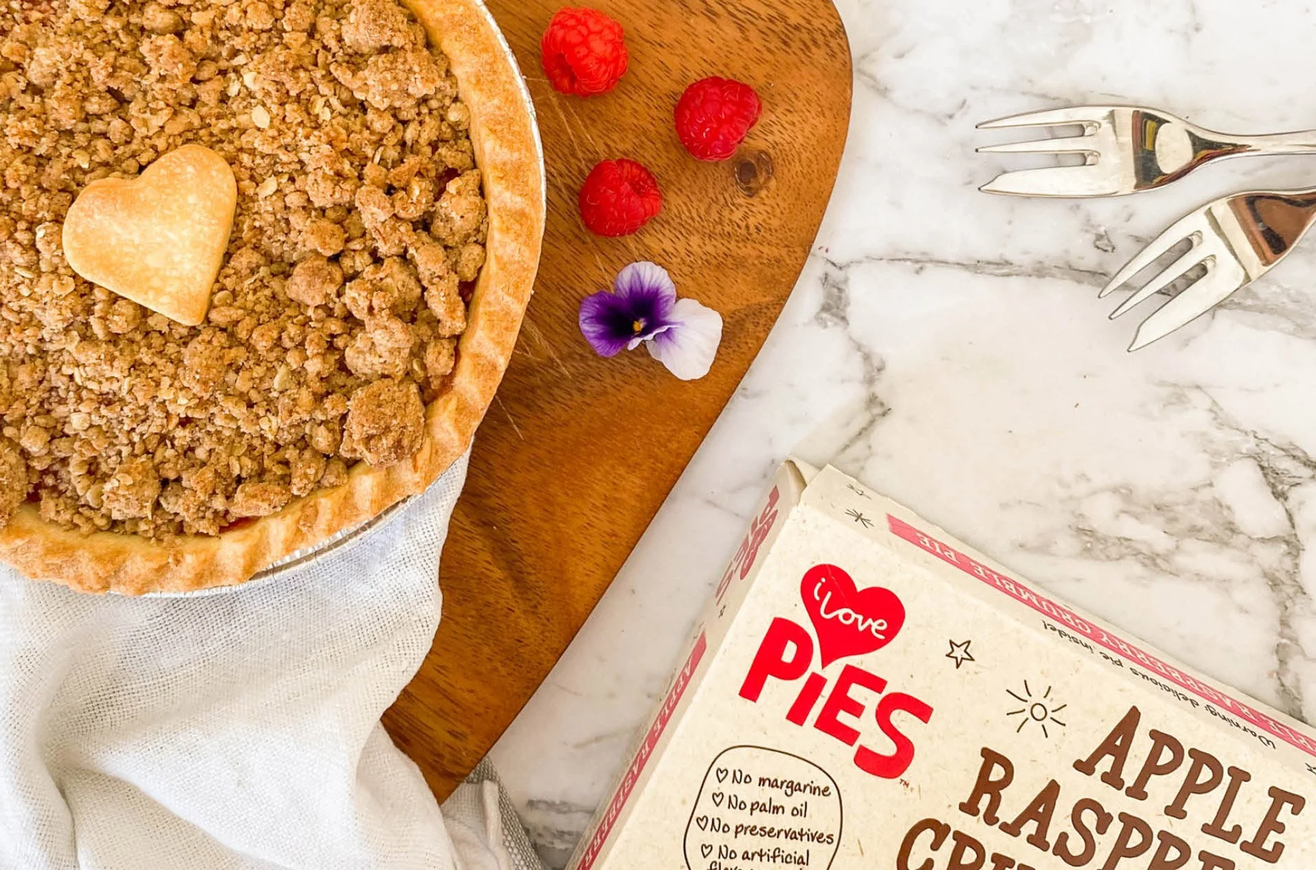 Social Media creative: A partially opened box of Love Pies Apple Raspberry Crisp with a crumbly topping, a heart-shaped cookie on the pie, a few raspberries, and an edible flower next to a wooden cutting board with two forks on a marble surface.