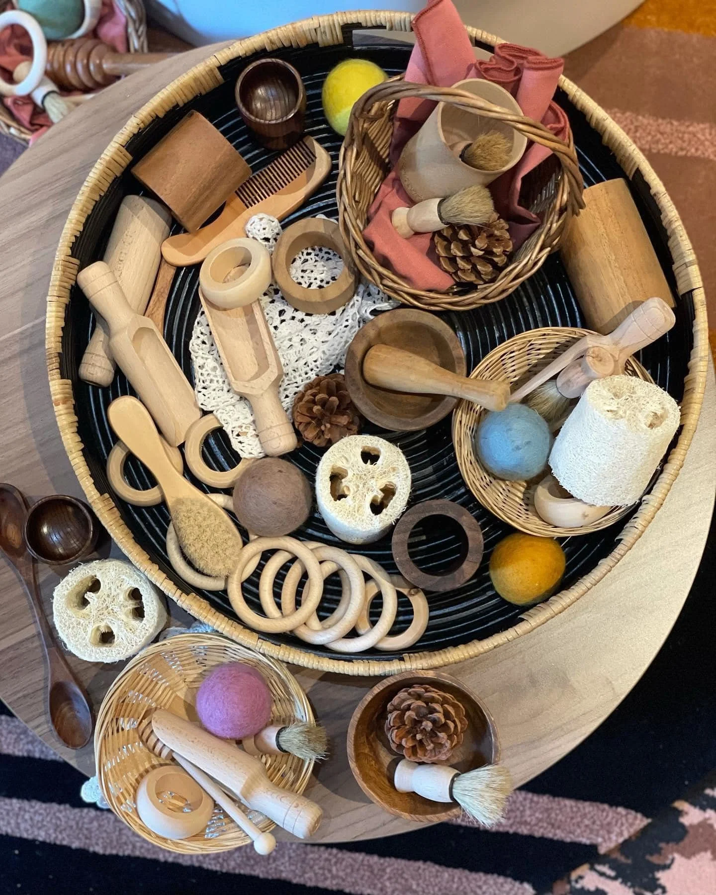 An assortment of wooden and natural fiber toys and objects, including spoons, rolling pins, circles, balls, pine cones, and sponges, arranged on a round black tray on a wooden table.