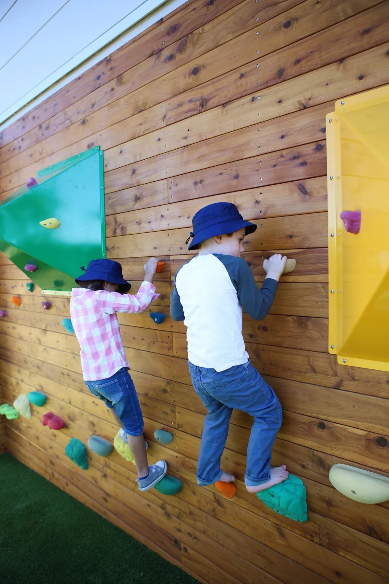 Two children wearing blue hats climbing on a wooden indoor rock climbing wall with colorful holds.