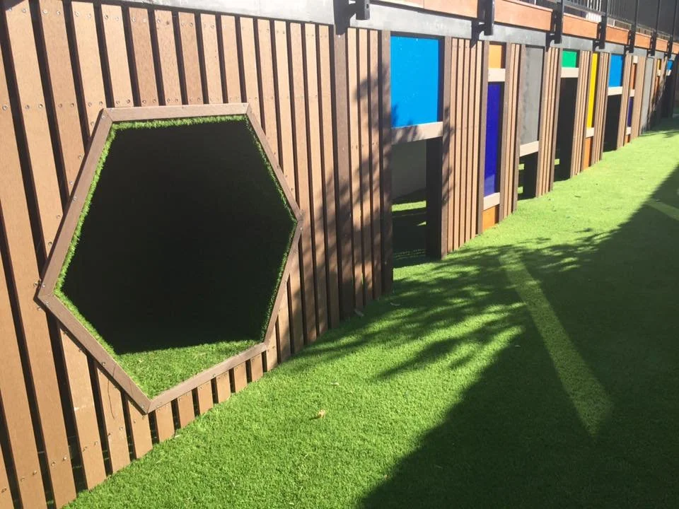 Colorful geometric window panels on a wooden fence with artificial grass on the ground and shadows cast by nearby trees.