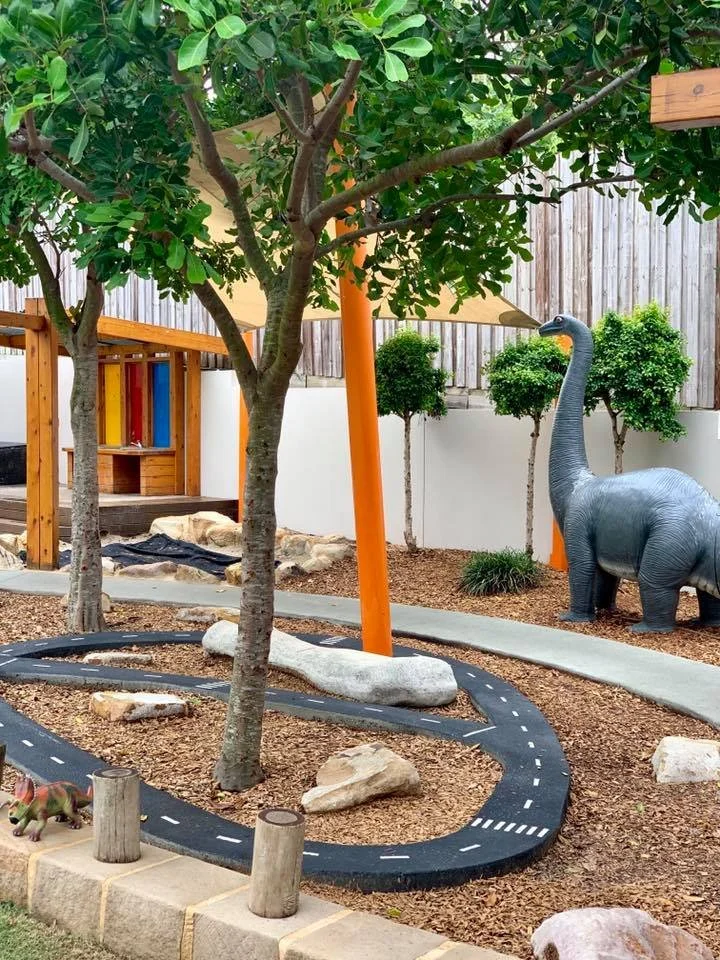 A miniature outdoor play area with a winding black rubber track, small trees, rocks, and a large plastic elephant statue.