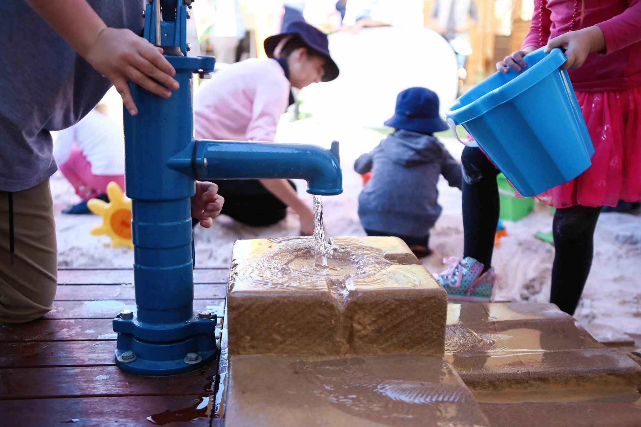 Children playing in a sandbox with a blue water fountain, water flowing onto sand, others using buckets, a woman supervising in the background.