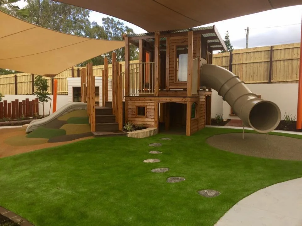 Backyard play area with a wooden playhouse, slides, and a grassy yard with stepping stones.