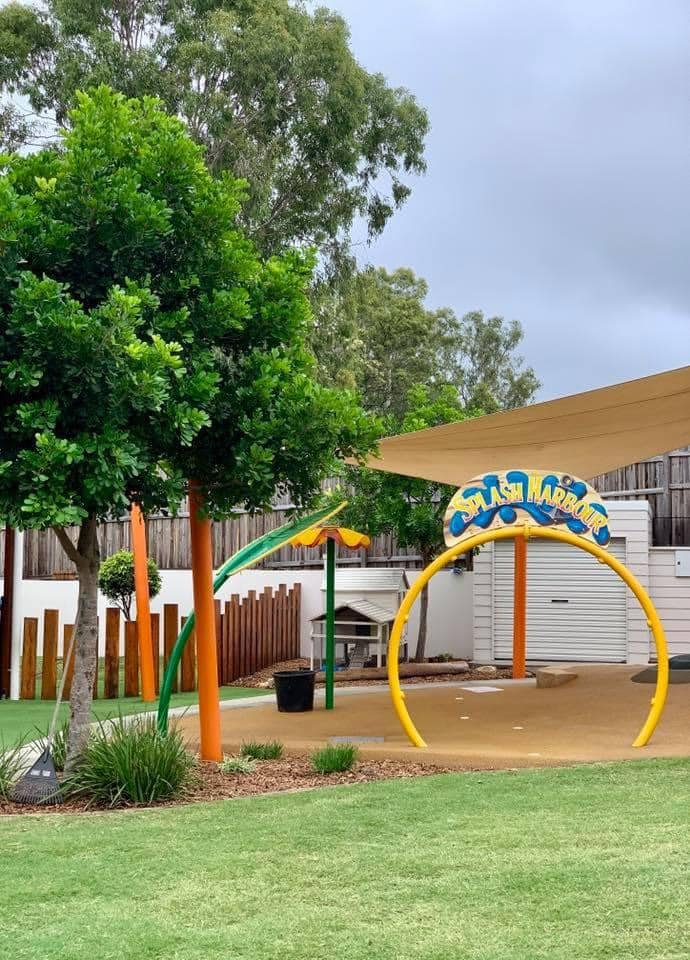 A splash playground area with green trees, a yellow climbing structure, and a colorful sign reading 'SPLASH HARBOUR'.