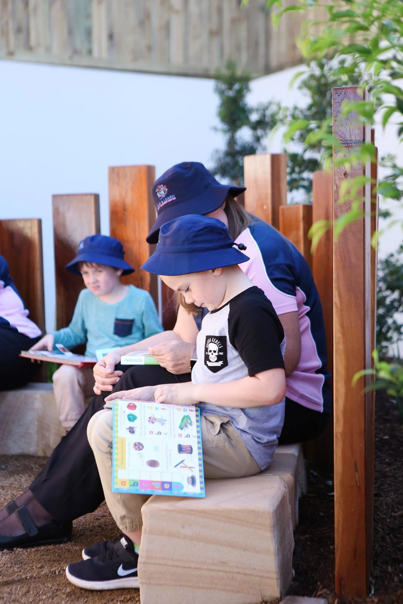 Children sitting outdoors on a stone bench, reading books, wearing blue hats, with a wooden fence and greenery behind them.