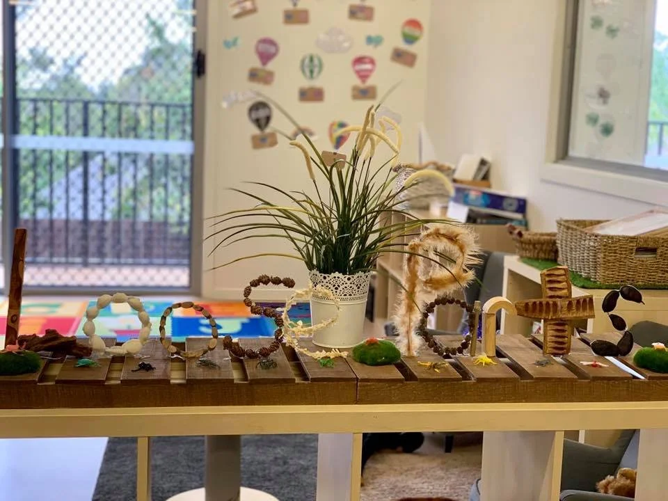 A table with various handmade craft decorations and a potted plant, situated in a well-lit room with a sliding glass door and furniture in the background.