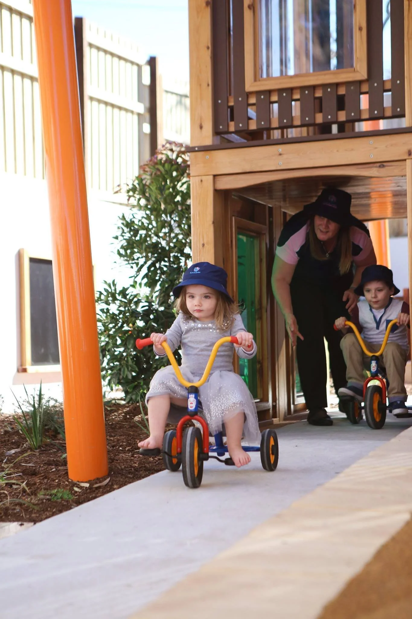 Young girl riding a tricycle outdoors with two children on similar tricycles and an adult woman assisting them near a wooden playhouse.