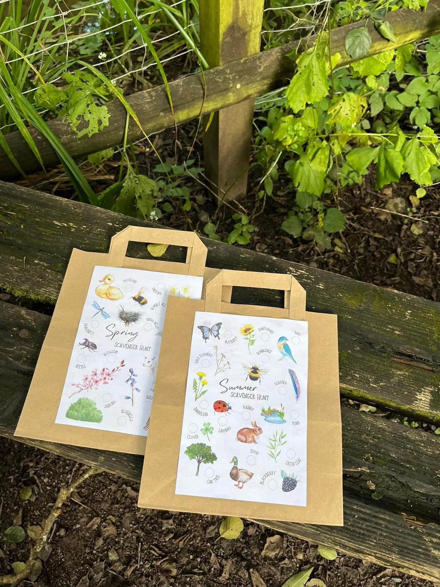 Two paper bags with outdoor seasonal scavenger hunt lists on them, placed on a weathered wooden bench outdoors among green plants and soil.
