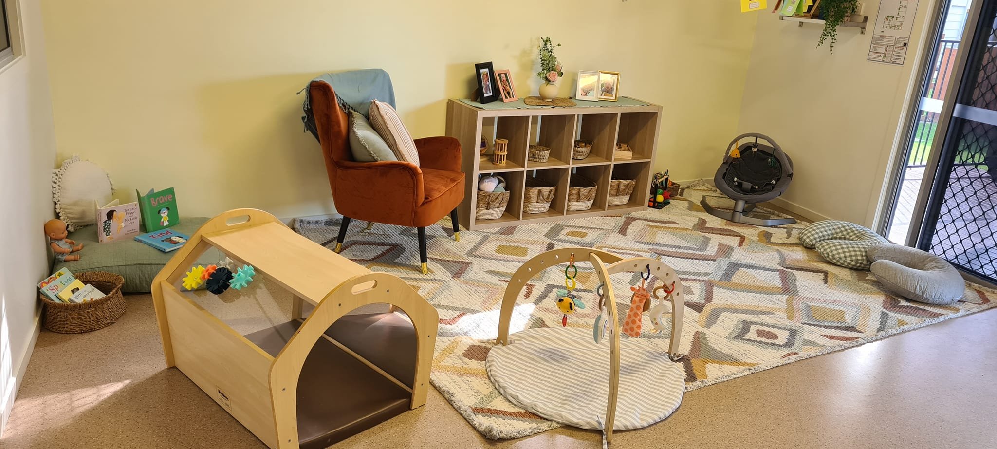 Playroom with a colorful geometric patterned rug, a brown shell rocker with pillows, a small wooden toy storage box, a cushioned floor seat with books and toys beside a basket, a wooden baby gym with hanging toys, a gray baby swing, and bed pillows n