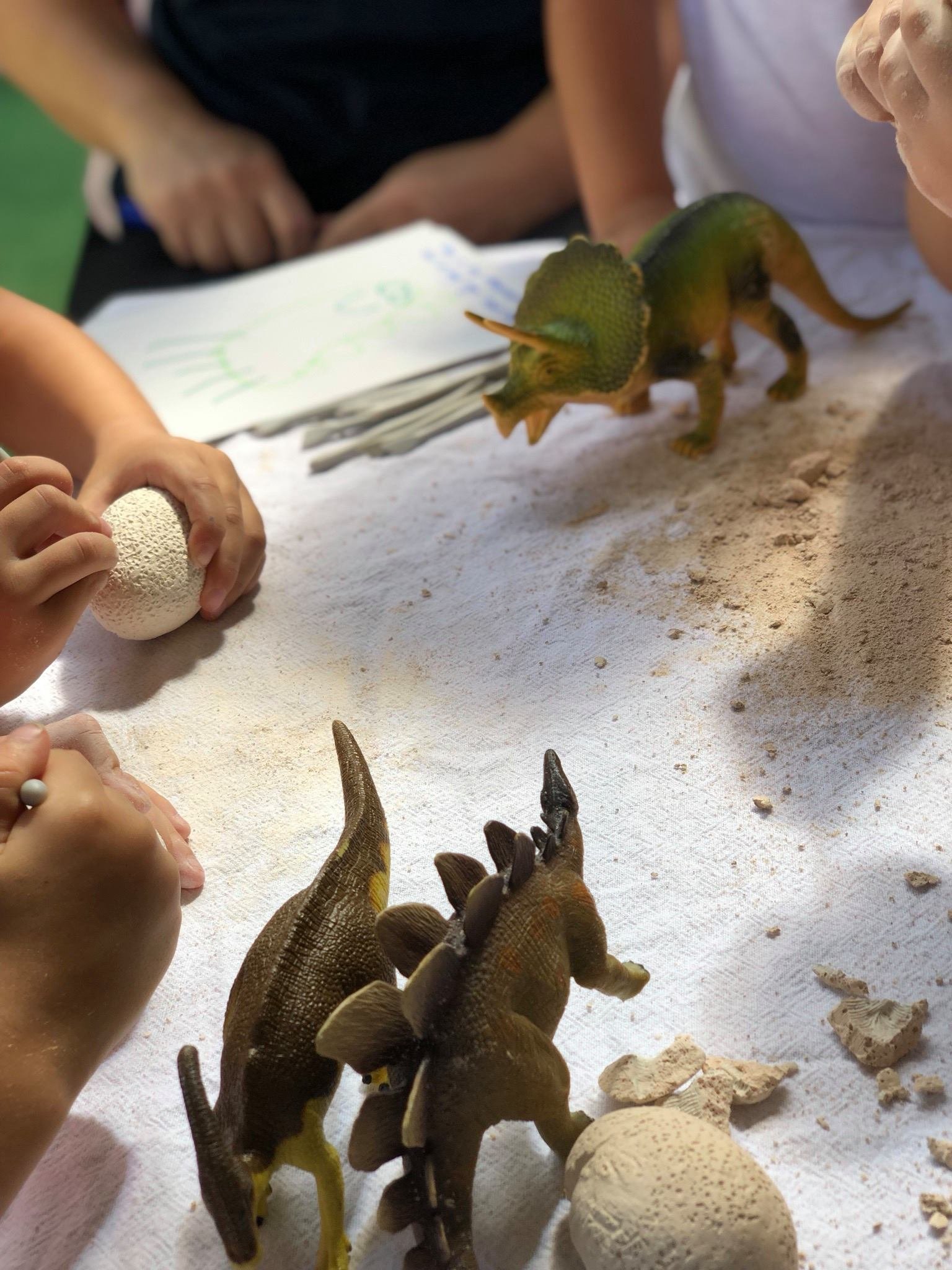 Children examining toy dinosaur models and a dinosaur figurine on a white surface with sand and an egg-shaped object.