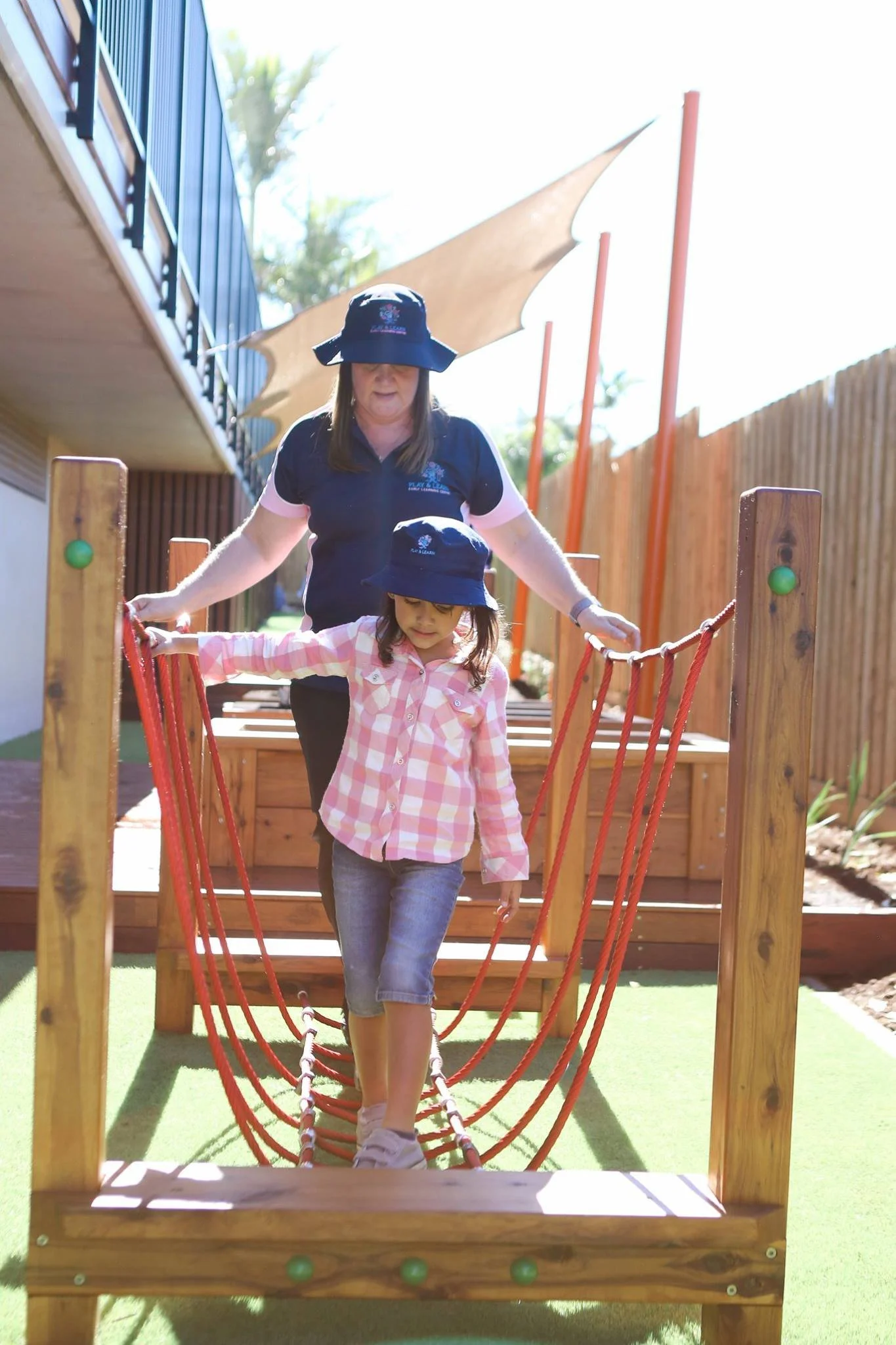 A young girl walks on a red rope bridge guided by an adult woman, both wearing dark blue hats, outdoors on a sunny day with a wooden fence and building nearby.