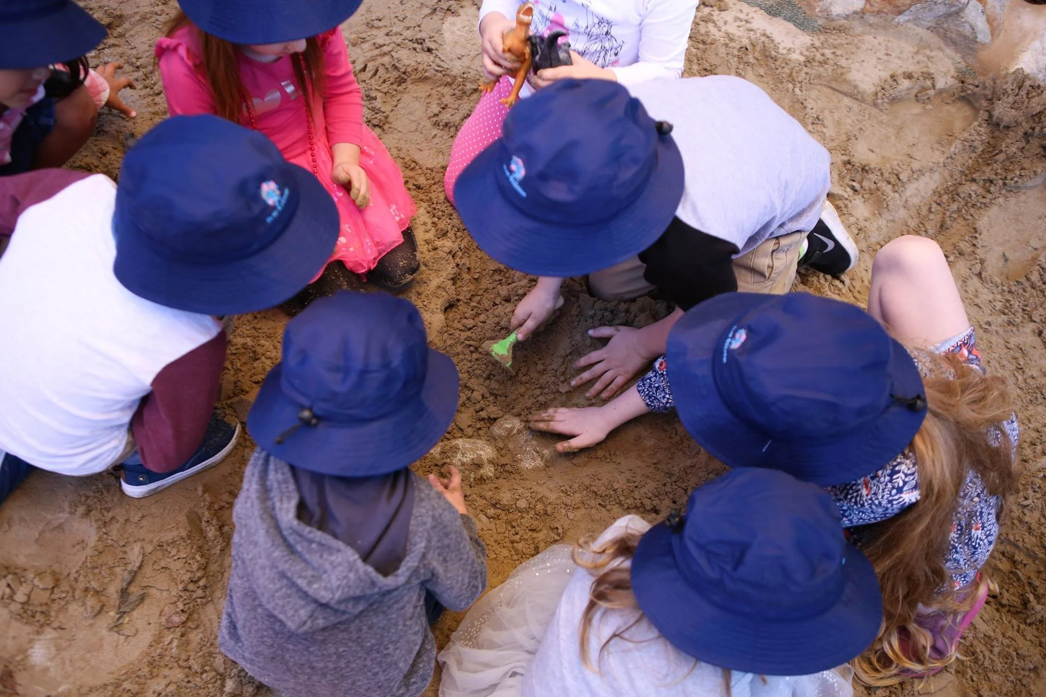 Group of children wearing blue sun hats playing in a sandbox, digging and using small green shovels.