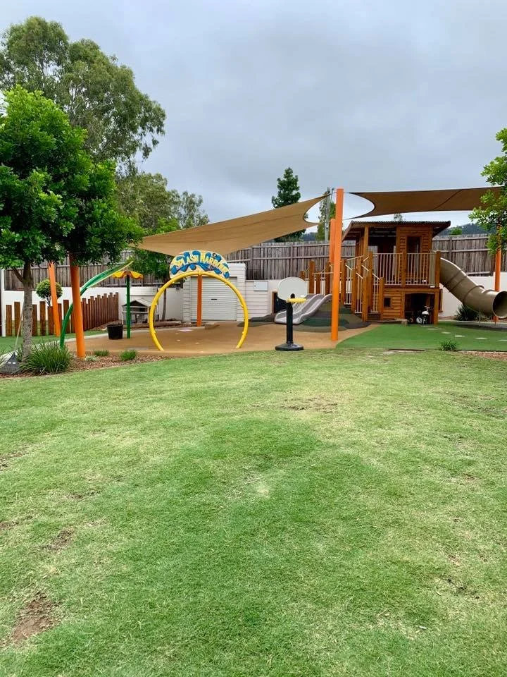 A playground with a wooden play structure, slides, a swing, and shade sails, surrounded by a grassy yard and trees.