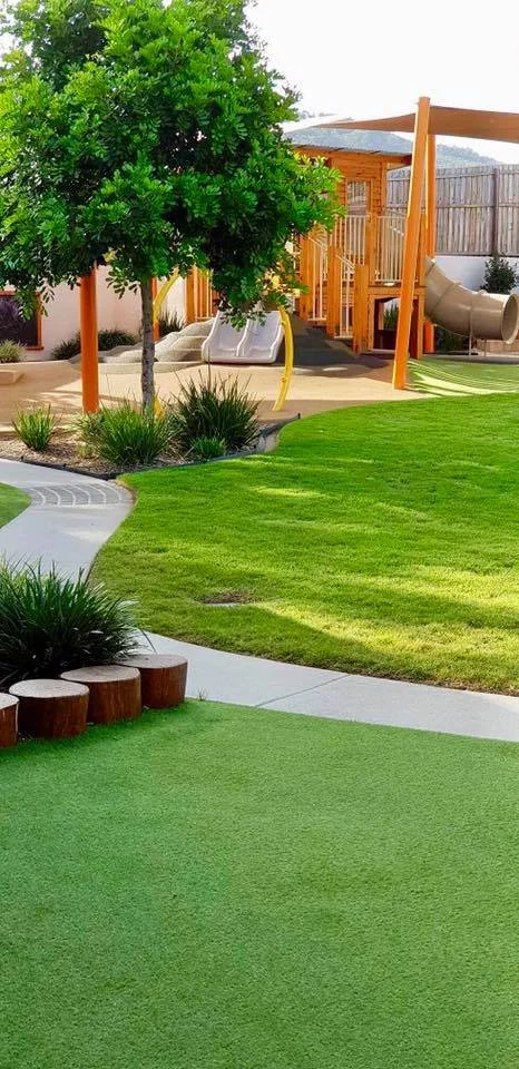 Colorful backyard with a grassy lawn, a large tree, a playground with a slide and a playhouse, and a winding concrete pathway.