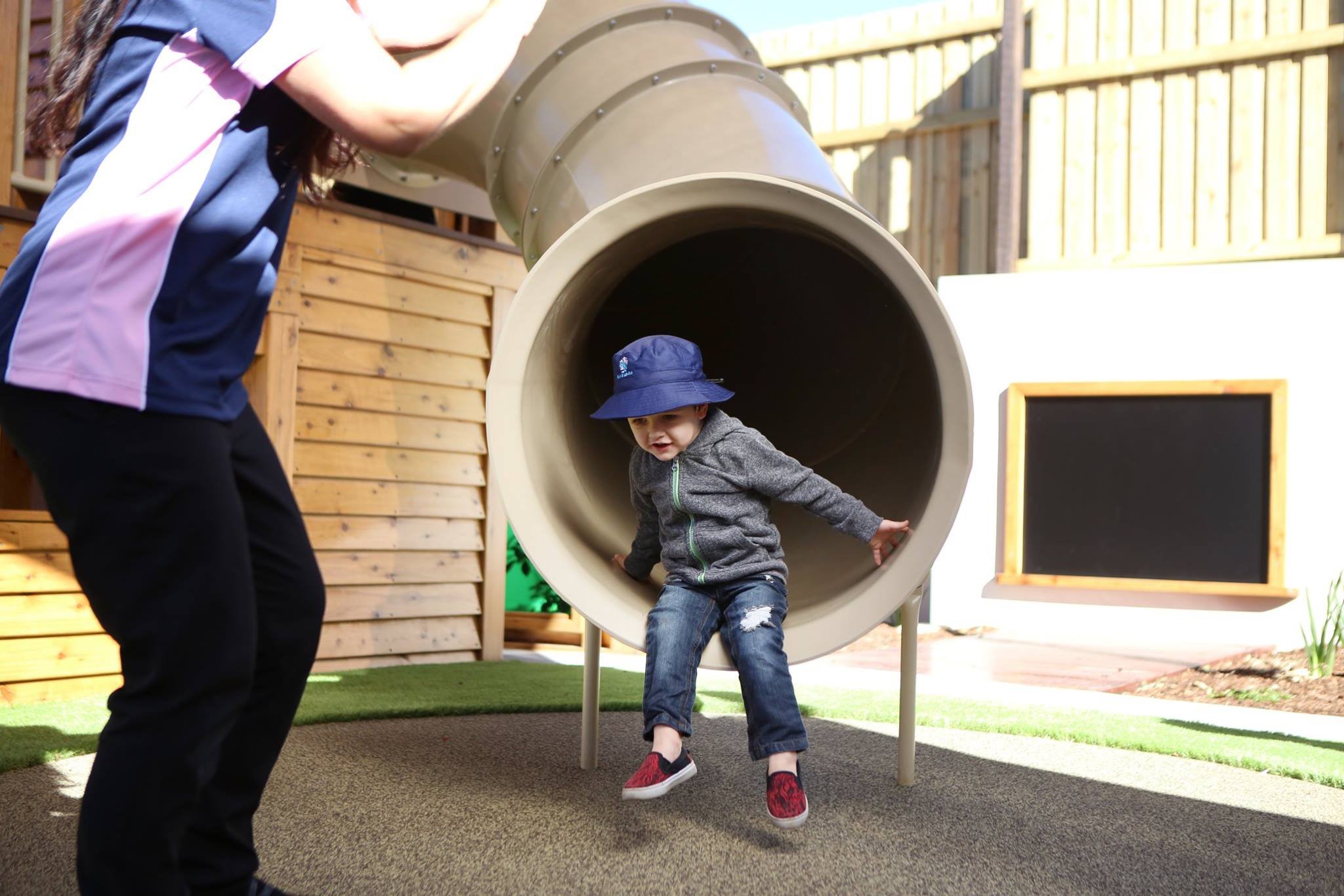 A young boy wearing a blue hat and gray hoodie is sitting at the entrance of a playground tunnel, with a person nearby in an outdoor play area with wooden fences and a small blackboard.