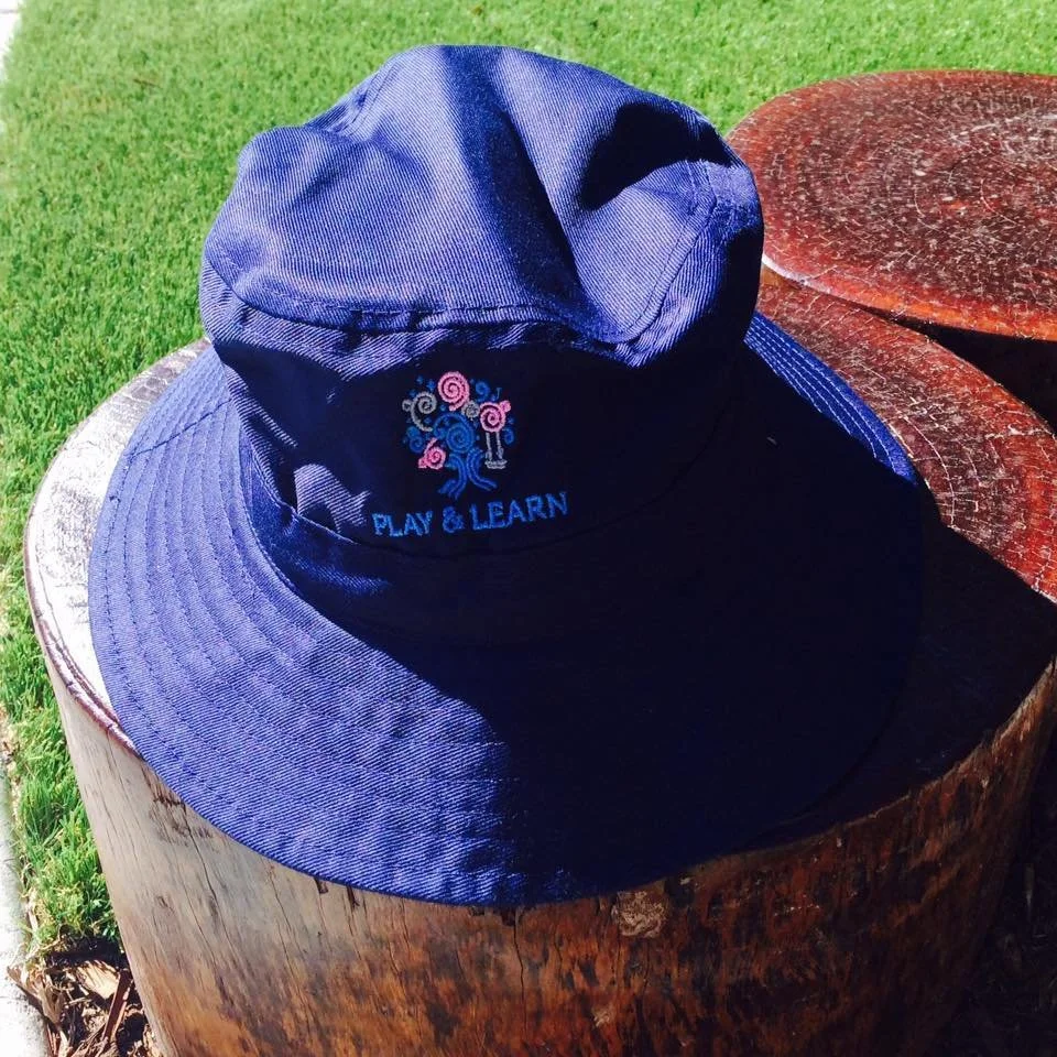 A navy blue bucket hat with embroidered colorful tree and the words 'PLAY & LEARN' on it placed on a wooden stump outdoors.