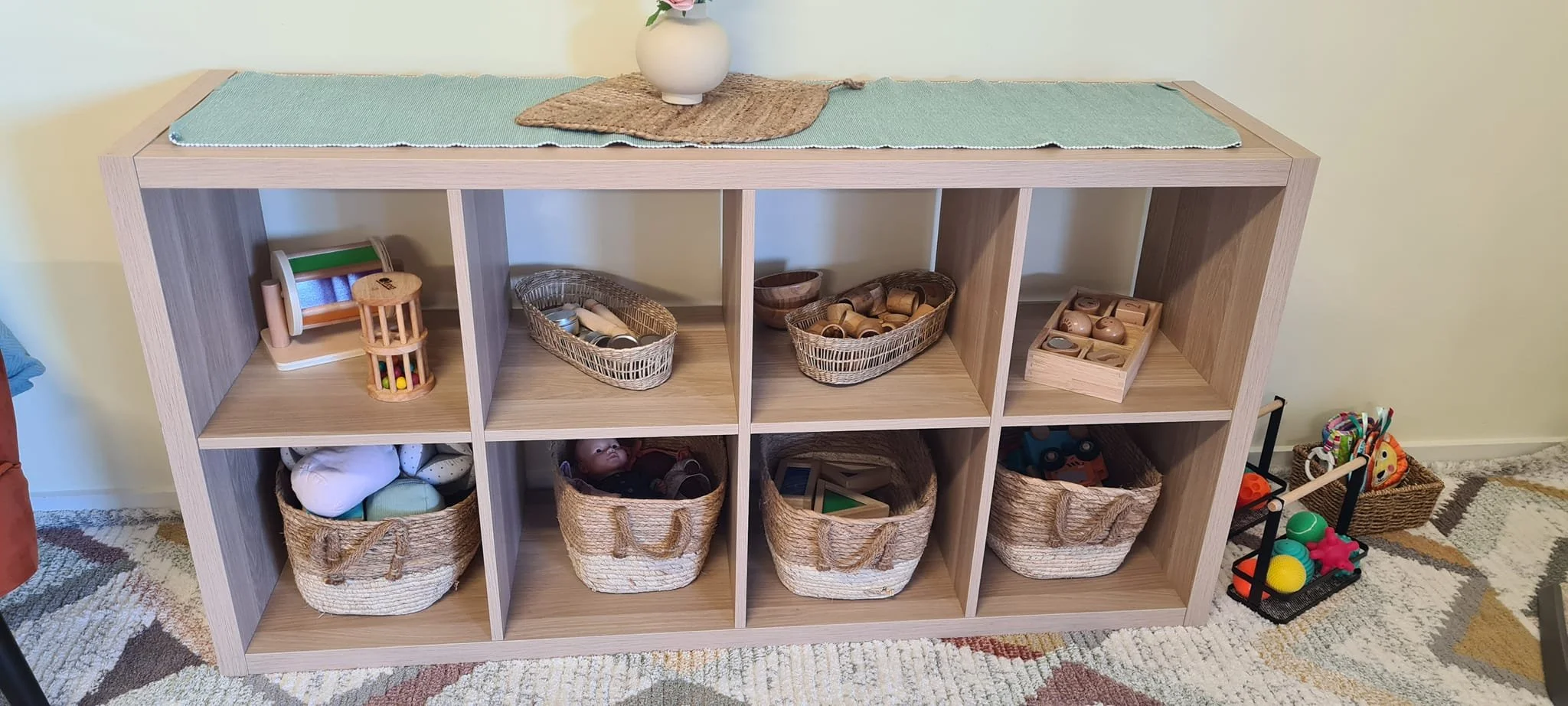 A wooden shelf with multiple compartments holding toys, baskets, and wooden blocks, with a small black and orange toy cart to the right and a colorful rug on the floor.