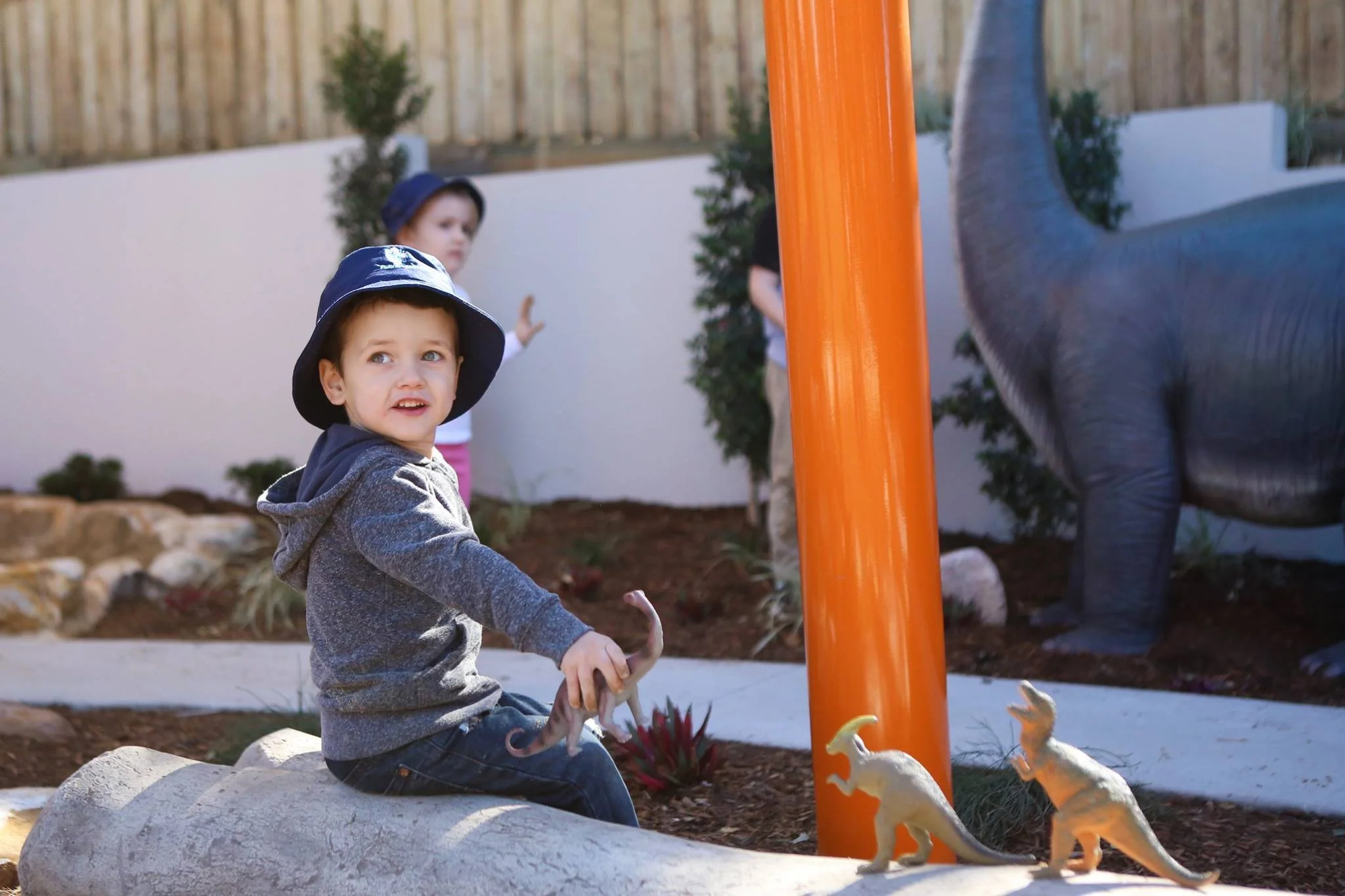 A young boy in a gray hoodie and black hat sitting on a log at a dinosaur-themed playground, holding a small dinosaur toy, with two other children and dinosaur statues in the background.