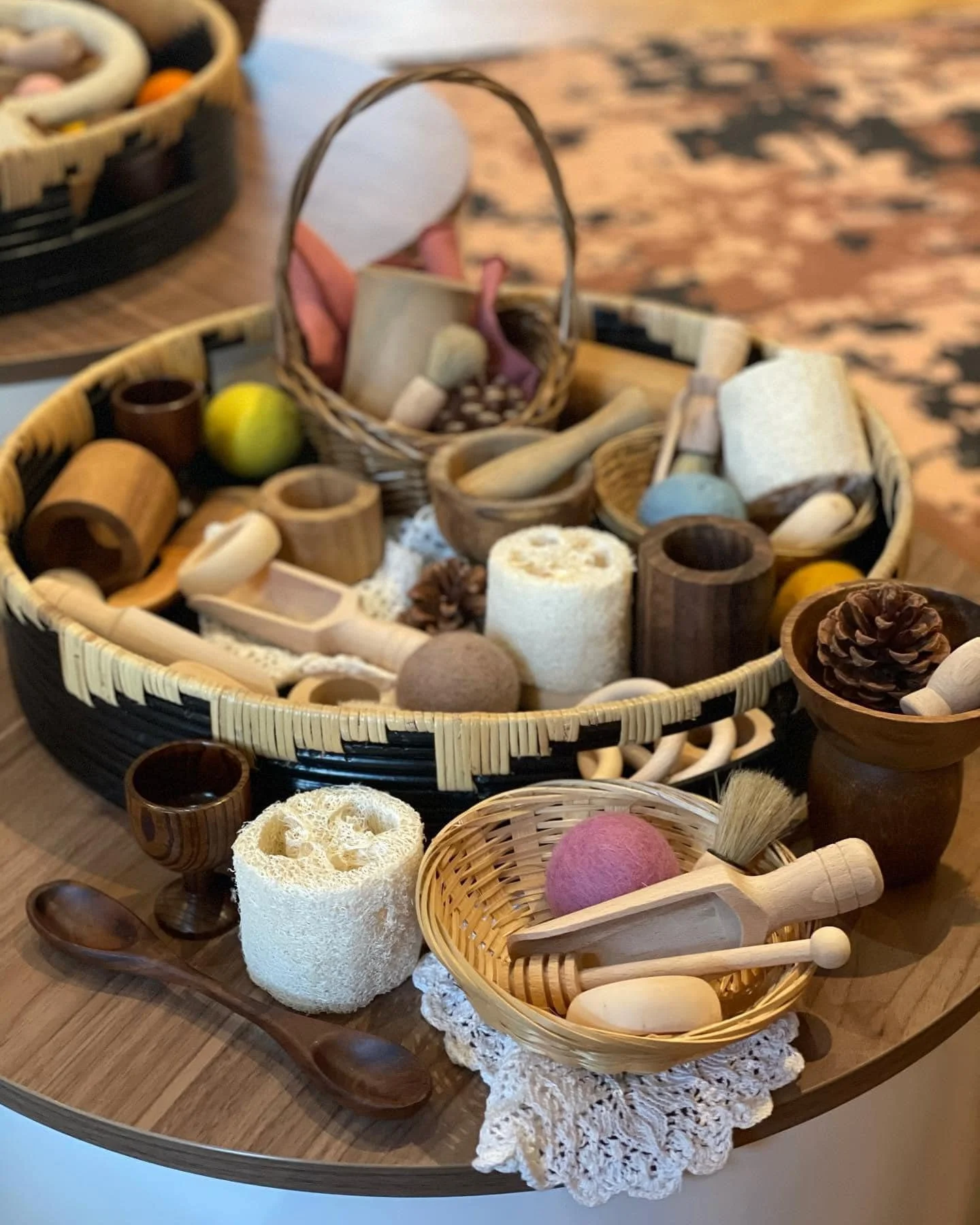 A collection of wooden and natural fiber bath accessories, toiletries, and decorative items in baskets on a wooden table, including loofahs, brushes, soap, pinecones, and small cups.