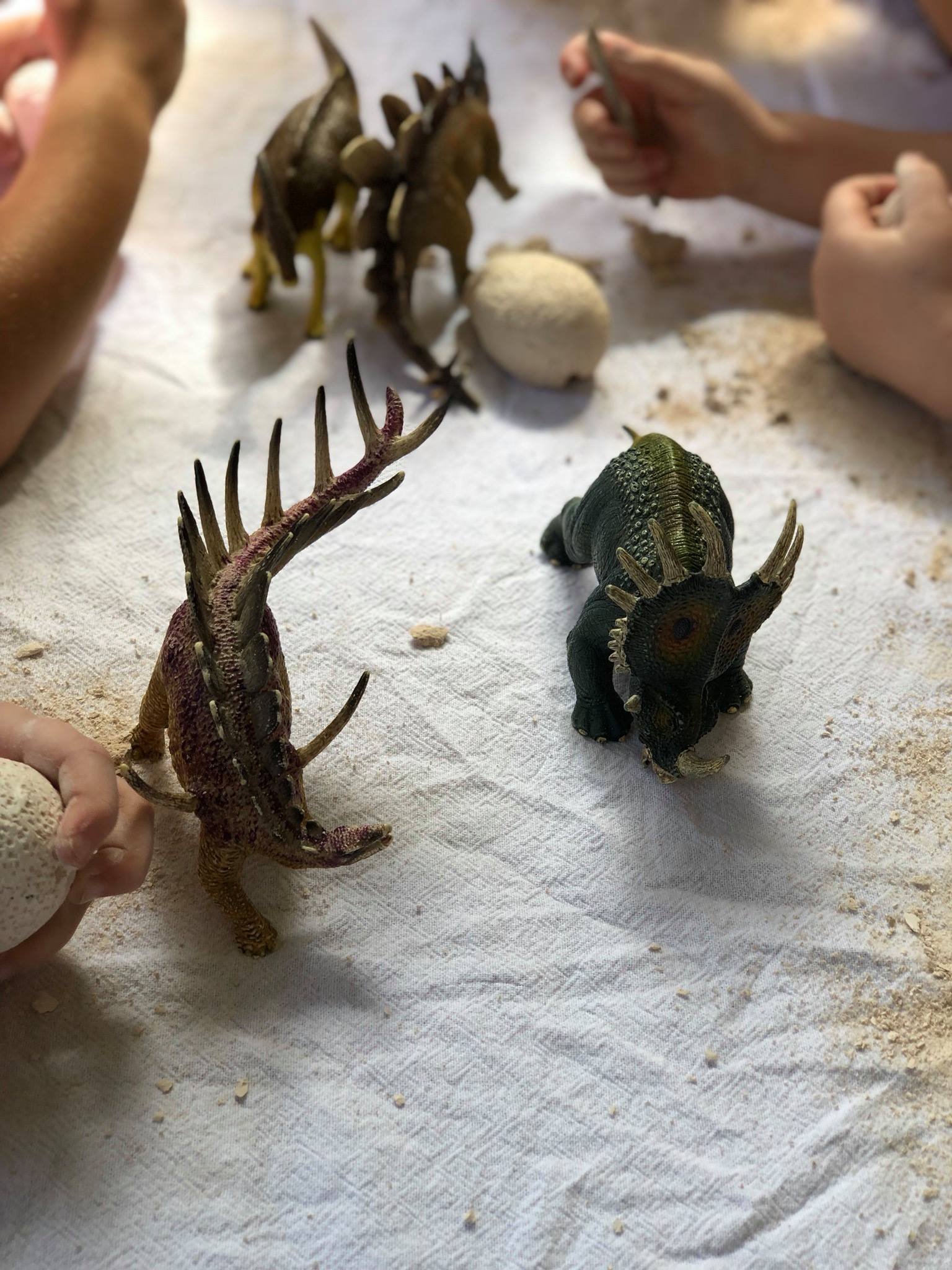 Children playing with toy dinosaurs and making sand or dirt balls on a white cloth surface.