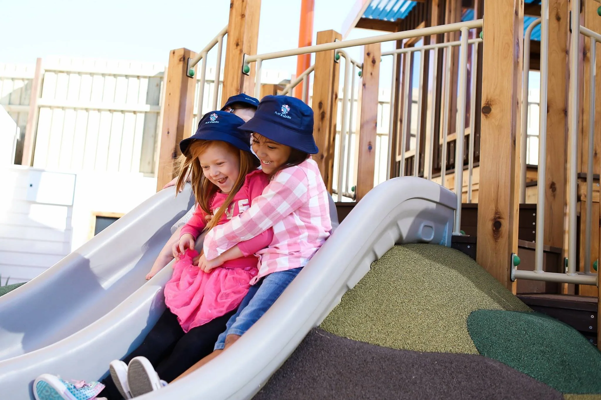 Two young girls wearing navy hats and pink clothing riding down a slide at a playground, smiling and enjoying themselves.