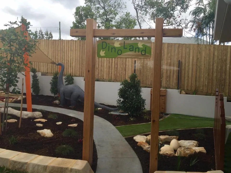 Backyard dinosaur themed play area with a life-sized dinosaur statue, a winding concrete path, small plants, a wooden fence, and a sign that reads 'Dino Land'.