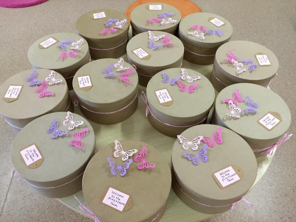 Several round gift boxes decorated with pink, purple, and white butterfly embellishments and small notes, arranged on a table, likely for a welcoming event.