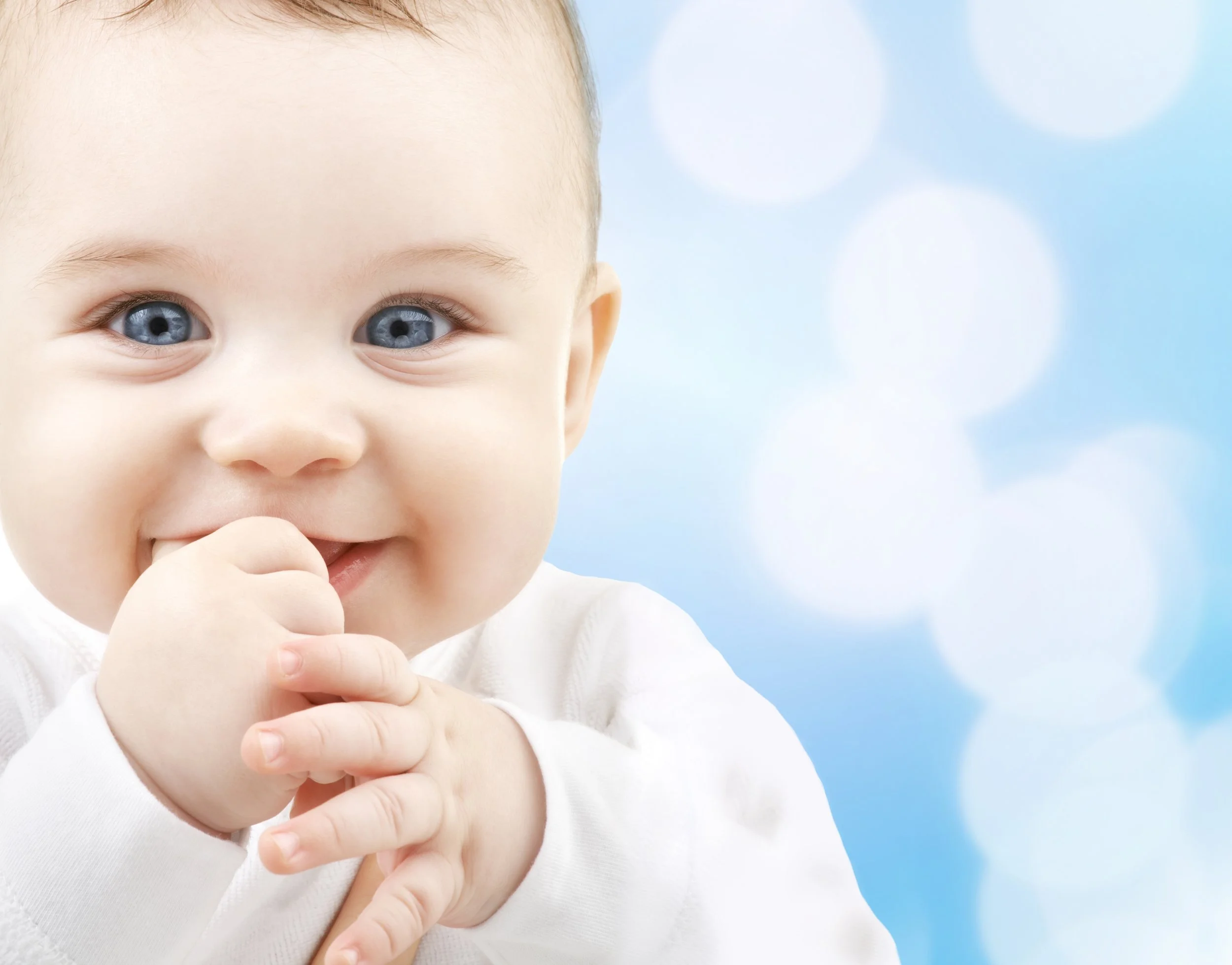 Close-up of a smiling baby with blue eyes, touching their mouth with one hand, against a soft focus blue background with bokeh.