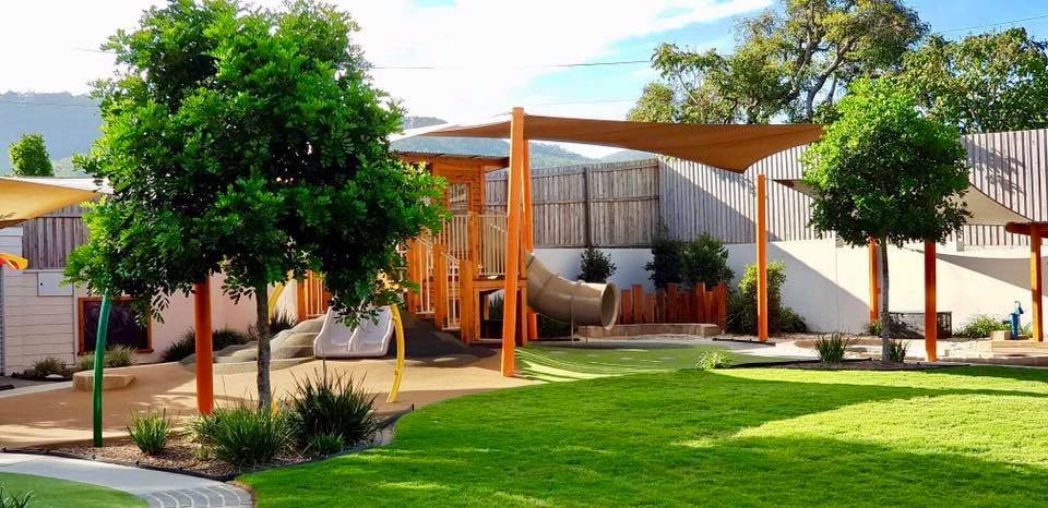 Playground area with slides, shade sails, trees, and a grassy lawn in a backyard