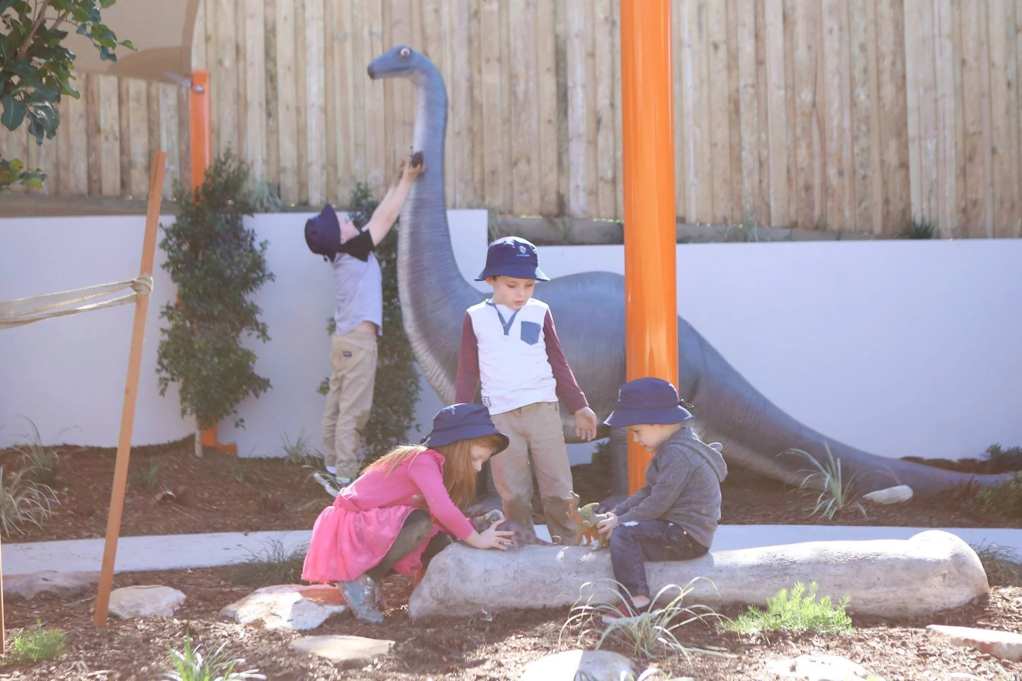 Children playing in a backyard garden with a dinosaur sculpture painted on the wall behind them. Four kids are wearing navy hats, and they are gathered around a log, playing with small dinosaur toys.