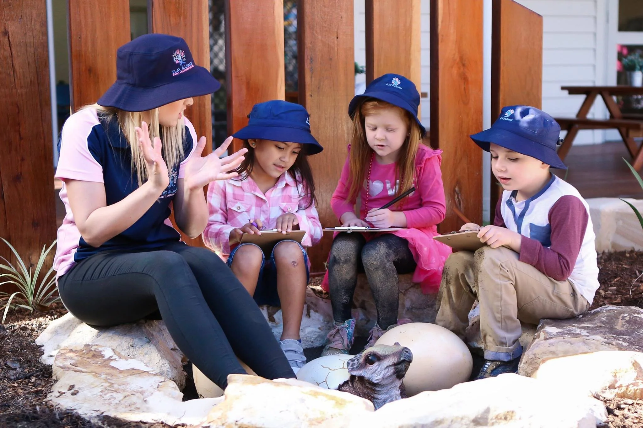 A teacher and four children sitting by a small outdoor pond with a baby alligator, examining and drawing in notebooks; the children are wearing blue hats and casual clothes.