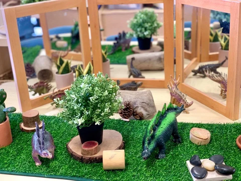 Miniature dinosaur figurines, potted plants, logs, and small stones arranged on fake grass with wooden frame and mirrors in the background creating a diorama scene.