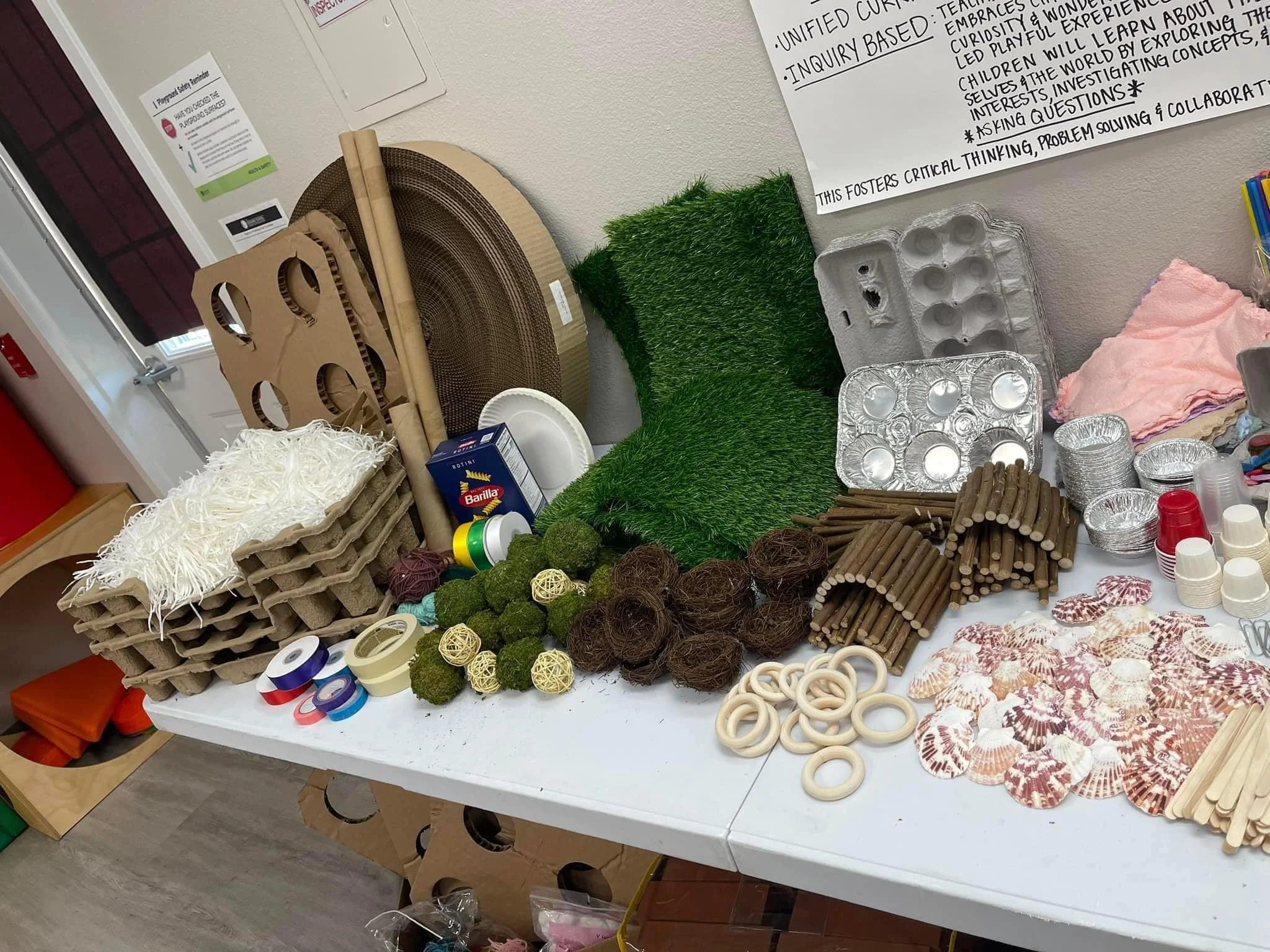 A table with various craft materials including cardboard, foam, moss, twine, seashells, and plastic cups, against a wall with a whiteboard and a door in the background.