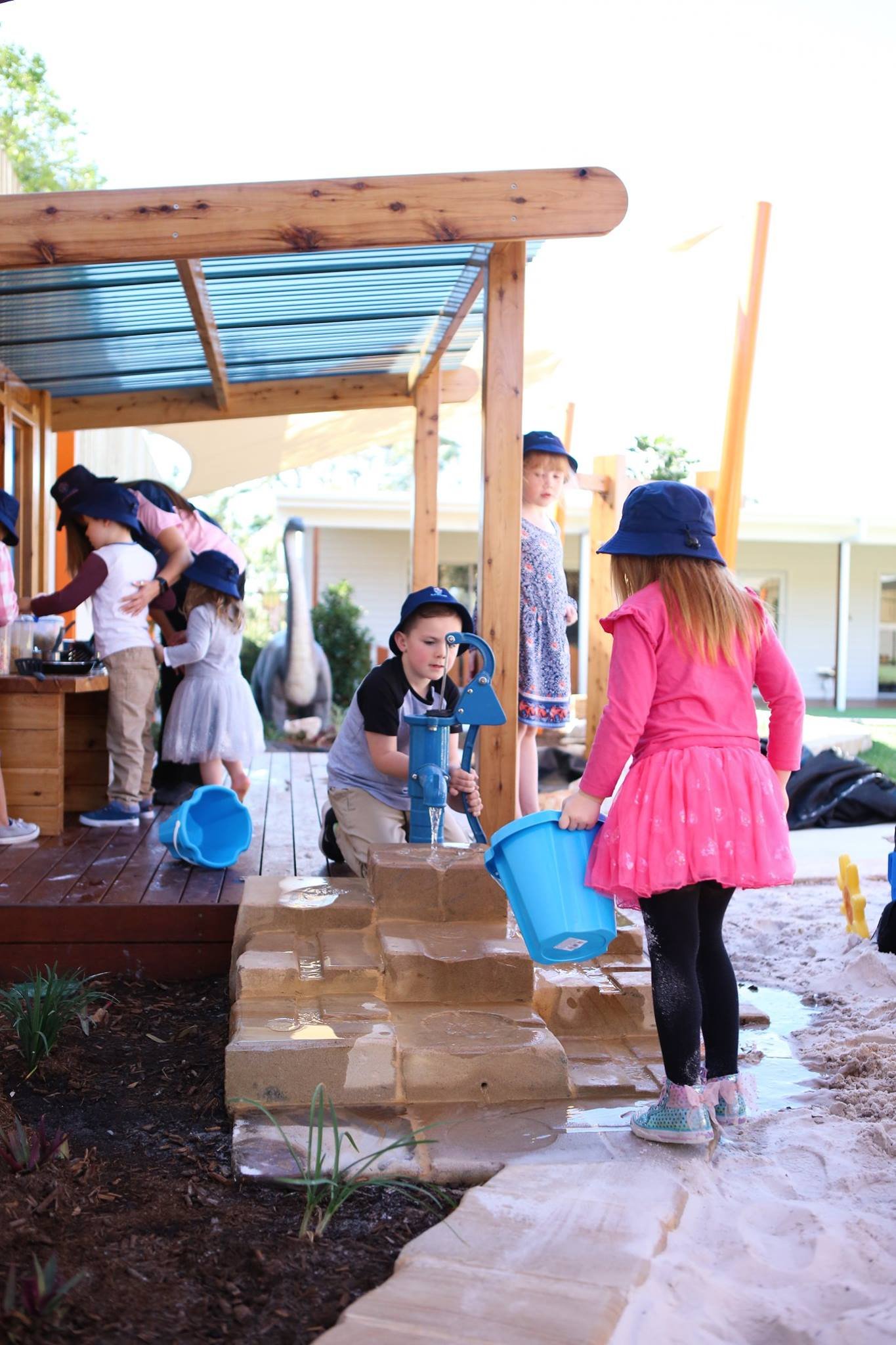 Children playing with water on a sand and stone play area at a playground with a wooden canopy.