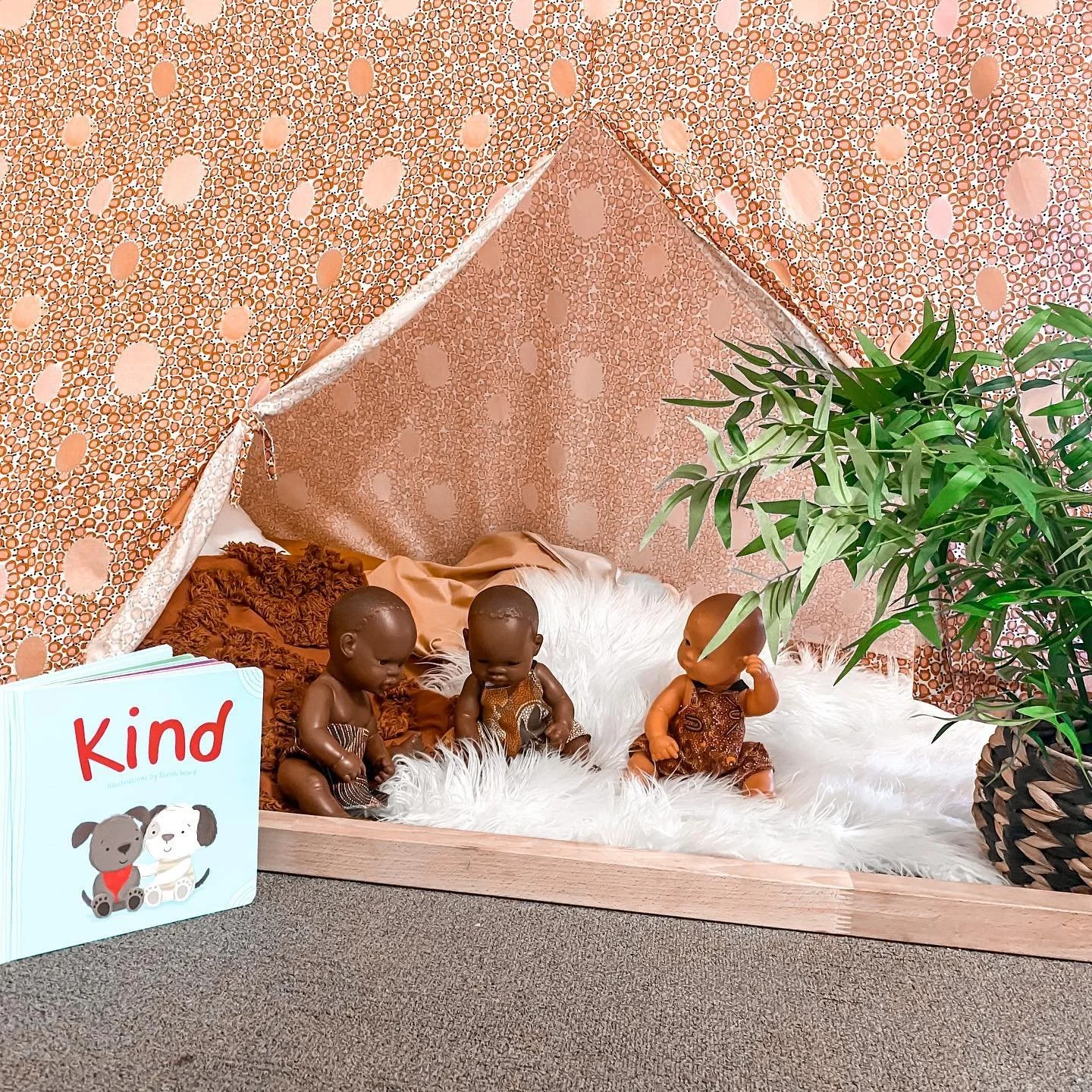 Three dolls sit on a fluffy white rug inside a small tent with pink and brown patterned fabric. A green plant is near the right side of the tent, and a book titled 'Kind' with illustrations of two puppies is on the floor outside the tent.